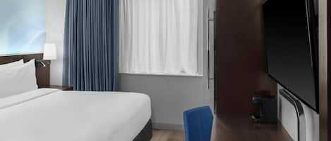 A hotel room with a bed, a desk with a flat-screen TV, a chair, a window with curtains, and a wall-mounted lamp.