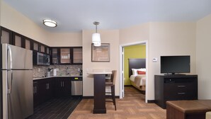 A compact hotel room with a kitchenette, a bed with a green accent wall, a television, and a small seating area. - Staybridge Suites Tomball - Spring Area by IHG (Tomball)
