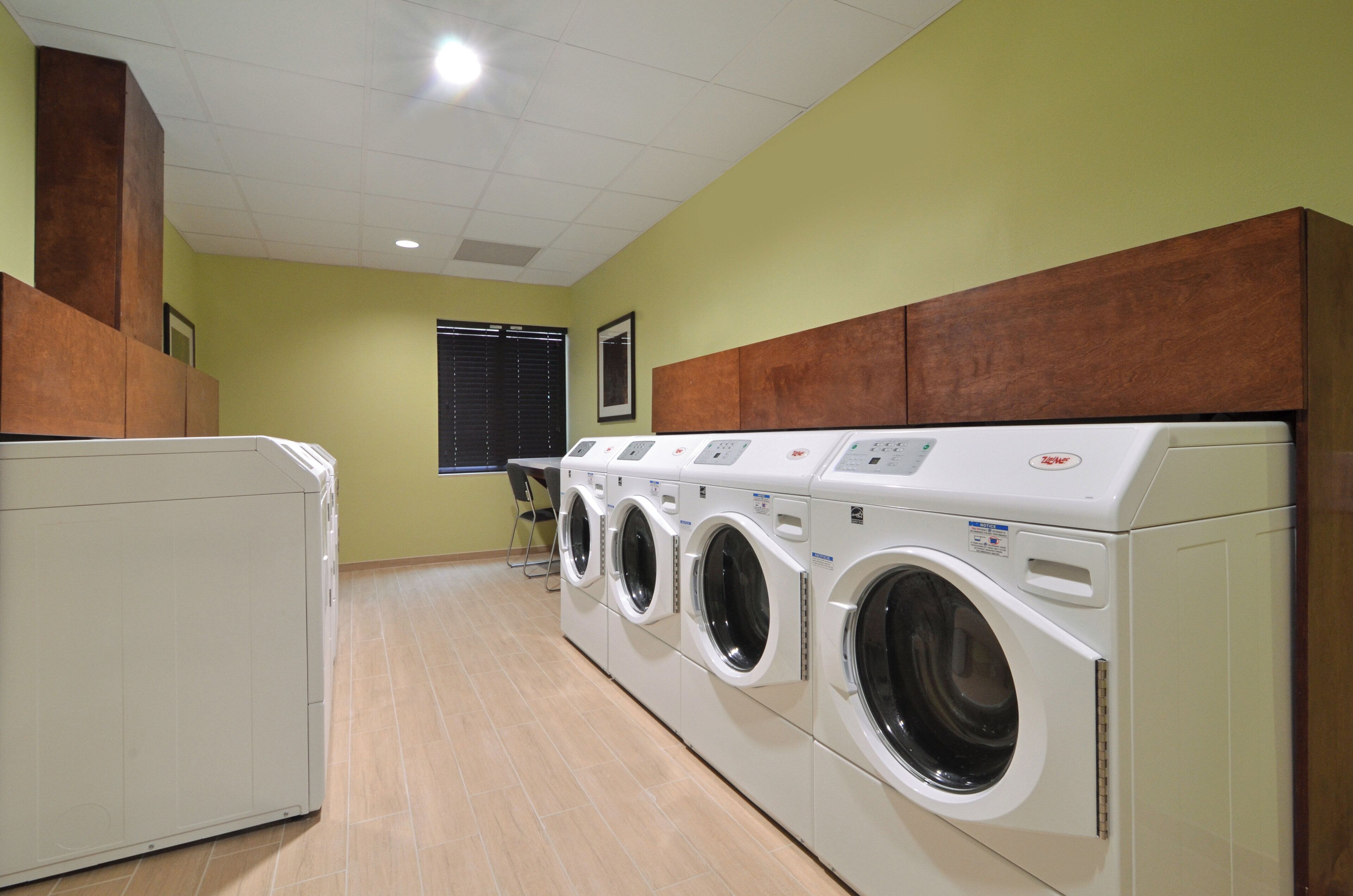 A laundry room with multiple washing machines and dryers, wooden cabinets above the dryers, and a window with blinds in the background.