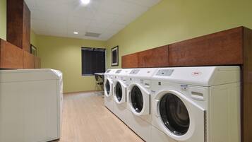A laundry room with multiple washing machines and dryers, wooden cabinets above the dryers, and a window with blinds in the background.