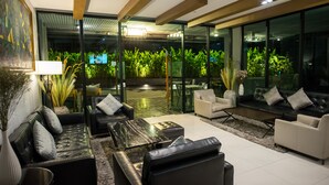 A modern living room with a glass wall, black leather sofas, a coffee table, and a view of indoor plants. - Sanae' Hotel Nimman (Chiang Mai)