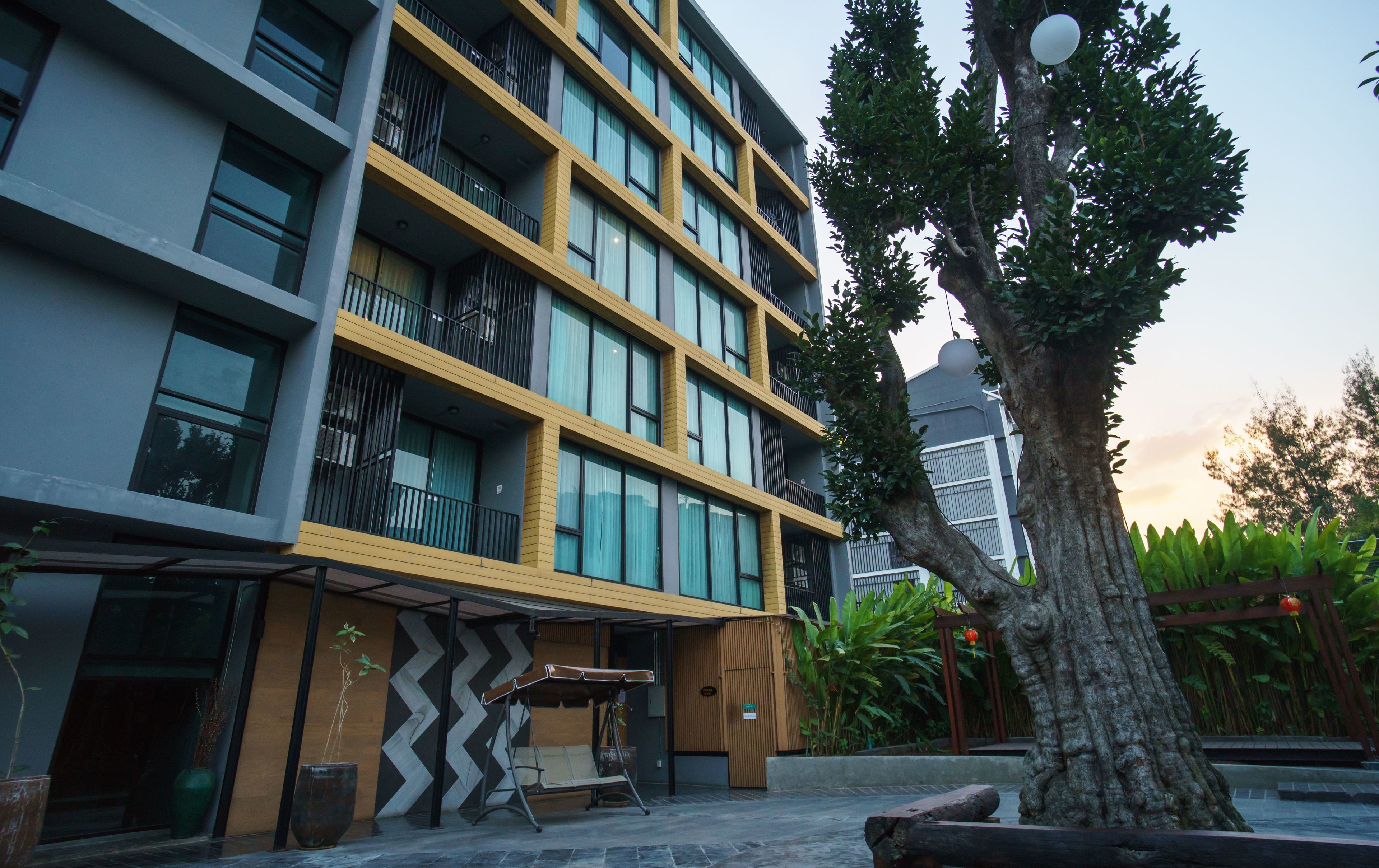 Un hotel moderno de varios pisos con balcones, una zona de estar al aire libre cubierta y un gran árbol en el patio.