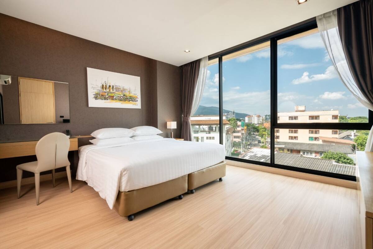 Une chambre d’hôtel avec un grand lit, un bureau, une chaise et une vue sur la ville grâce à de grandes fenêtres.