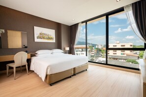 Habitación de hotel con una cama grande, un escritorio, una silla y vistas a la ciudad a través de amplias ventanas.