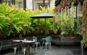 Un área de estar al aire libre con sillas de metal y una mesa, rodeada de plantas verdes exuberantes y una decoración colgante de plantas.