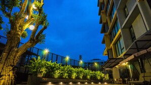 A nighttime view of a courtyard with a tree, potted plants, and a modern building with balconies. - Sanae' Hotel Nimman (Chiang Mai)