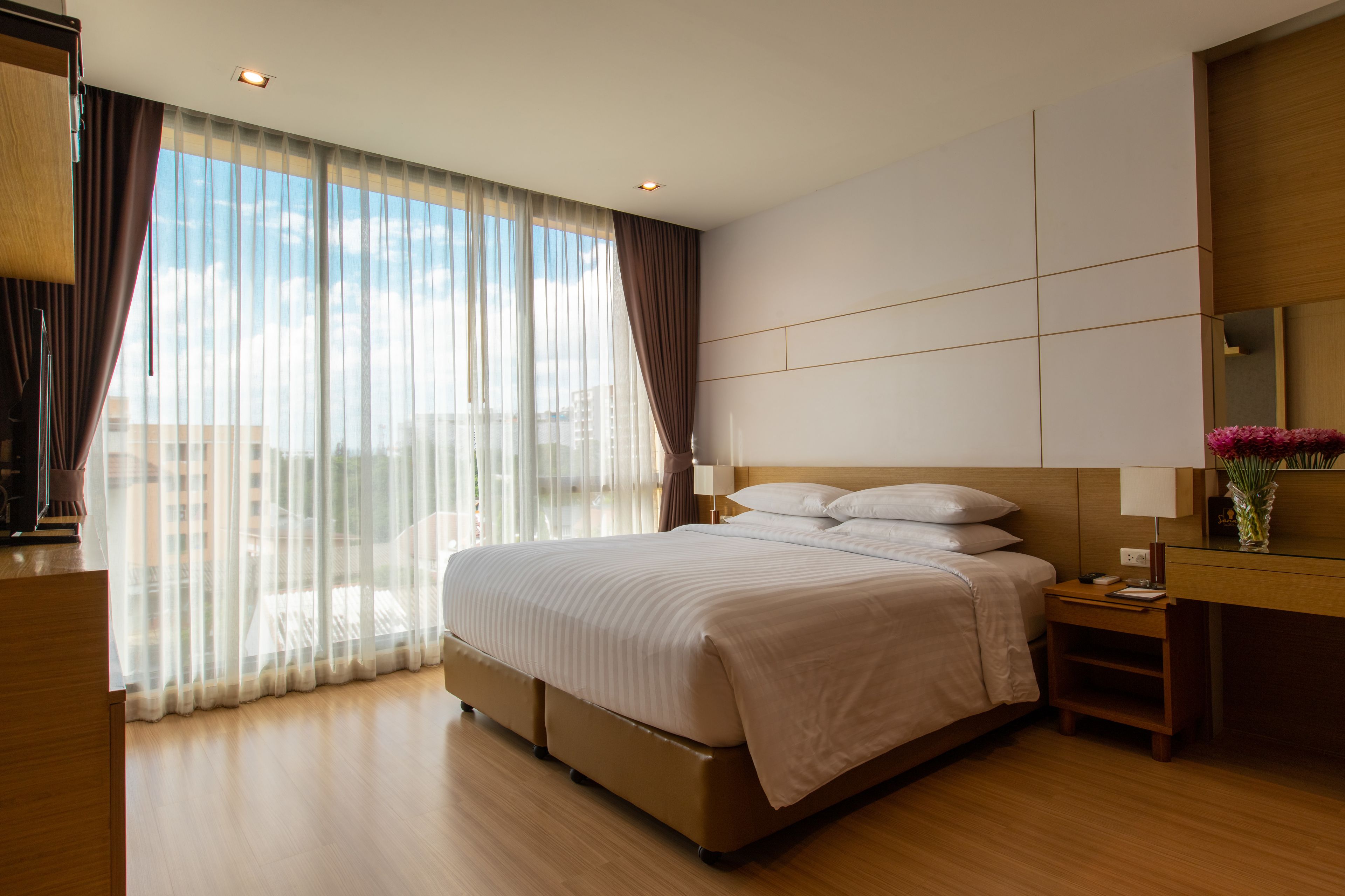 A modern hotel room with a large bed, a nightstand, a desk with a lamp, and a view of the city through sheer curtains.