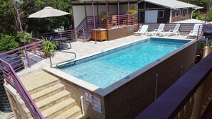 A rectangular pool with steps leading up to it, surrounded by a purple railing. There are lounge chairs and umbrellas around the pool area. -  JB'S SERENITY STAY (Carnbee)