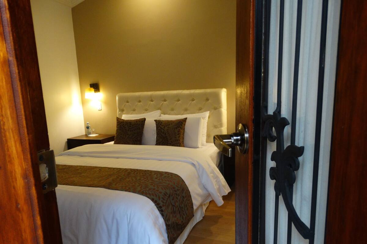 Ein Hotelzimmer mit einem großen Bett, einem Nachttisch, einer an der Wand befestigten Lampe und einer Tür mit einem dekorativen Griff.
