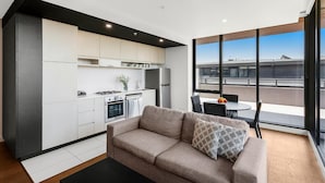A modern kitchen with white cabinets, a built-in oven, and a refrigerator. There is a dining area with a table and chairs, and a large window offering a view of the city. - Clayton Serviced Apartments (Clayton)