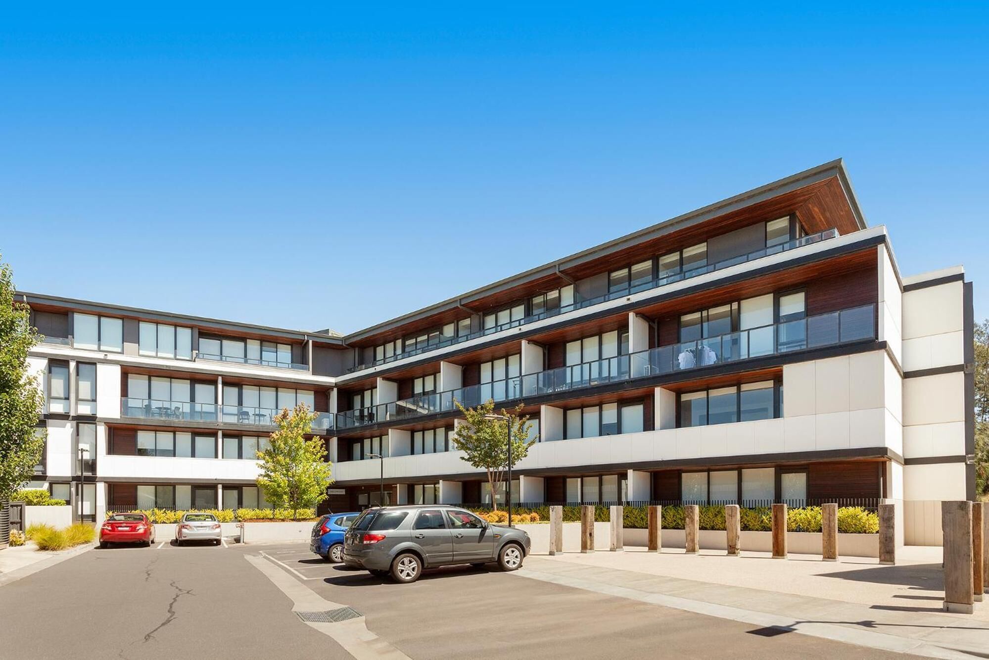 A modern multi-story building with balconies, a parking area, and a clear blue sky.