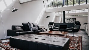 A modern living room with a sofa, armchair, ottoman, and coffee table. There is a large window providing natural light and a view of trees. - Alkina Lodge (Wattle Hill)