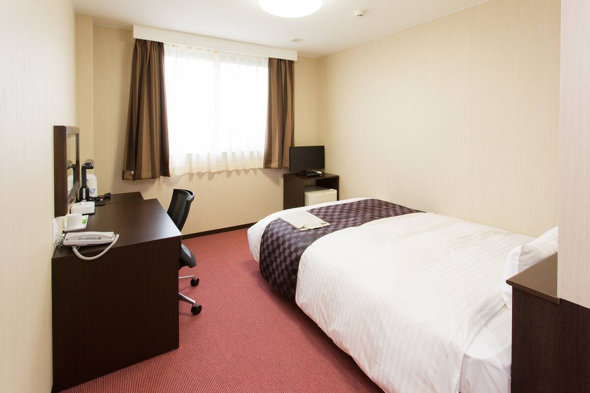 A hotel room with a desk, chair, two beds, a window with curtains, and a flat-screen TV.