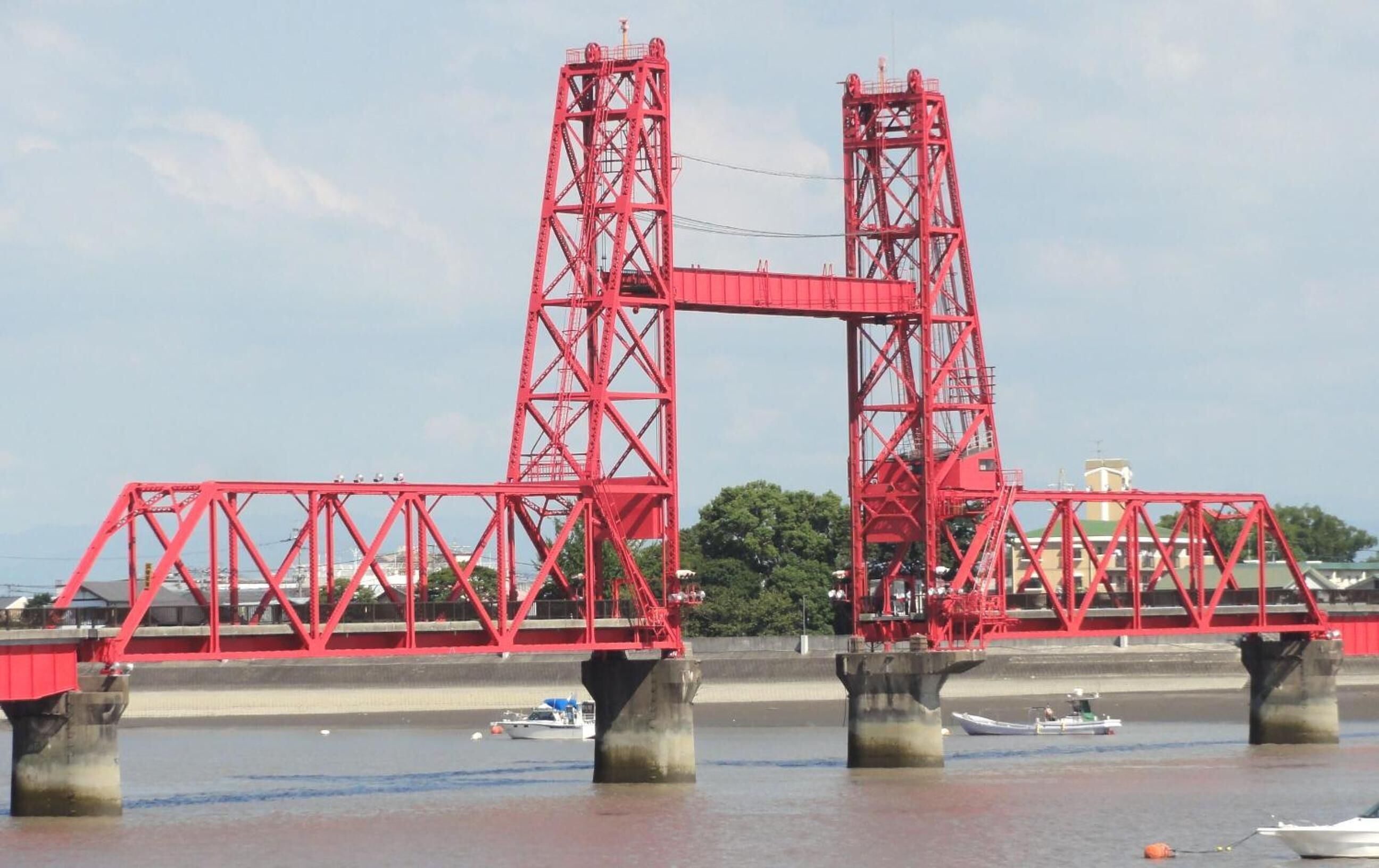 A red bridge with two towers, supporting a wide roadway over a body of water. Boats are visible on the water below.