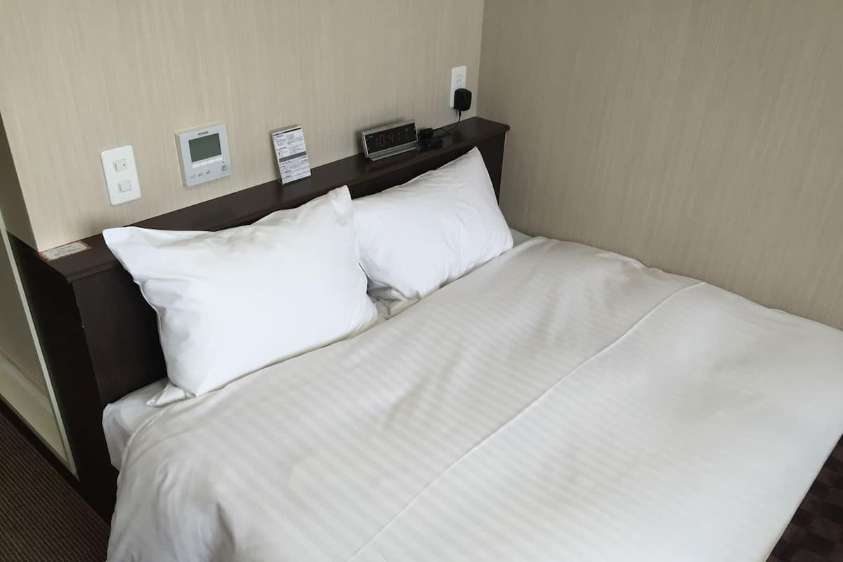 A hotel room with a bed, a control panel, and a digital clock on the wall.