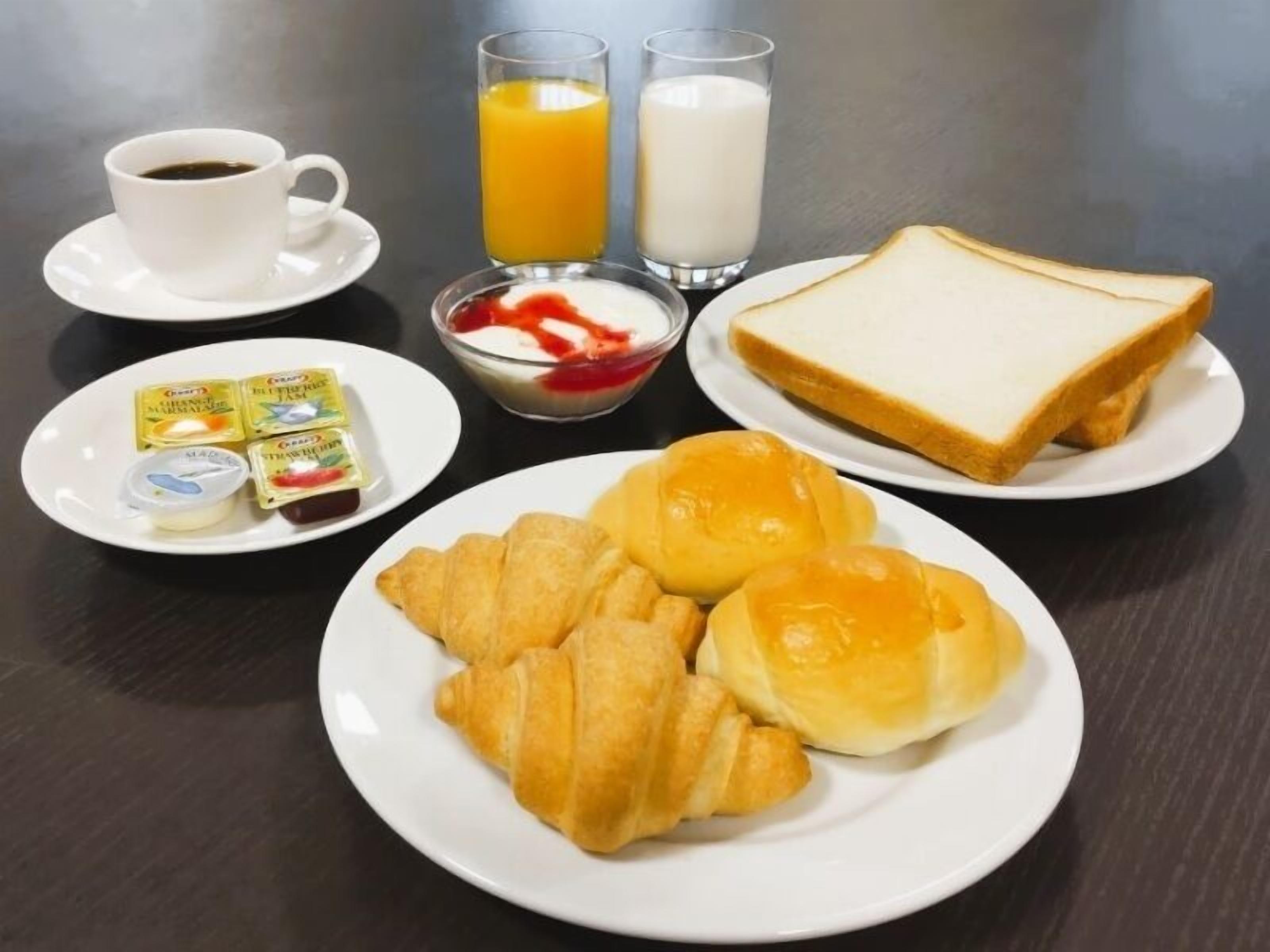 Una mesa de desayuno con croissants, pan, una taza de café, jugo de naranja, leche y varias opciones para untar en los platos.