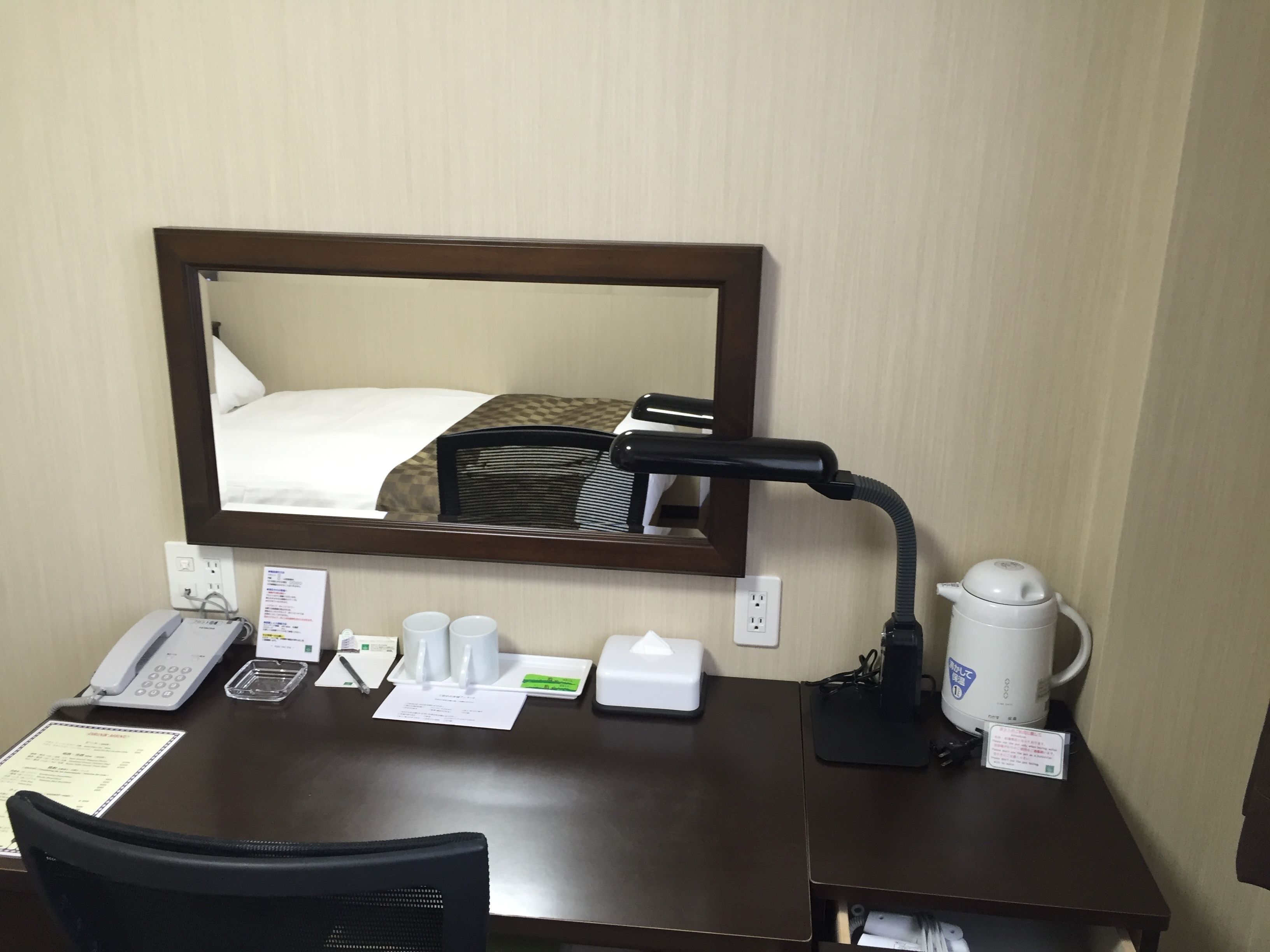 A hotel room desk with a telephone, a coffee maker, a mirror, and a bed with a patterned bedspread.