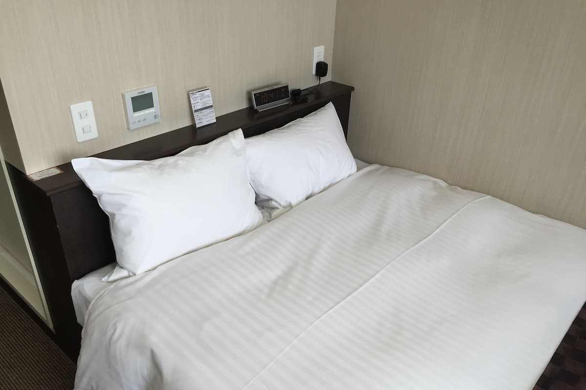 A hotel room with a neatly made bed, a digital clock, a thermostat, and a power outlet on the wall.