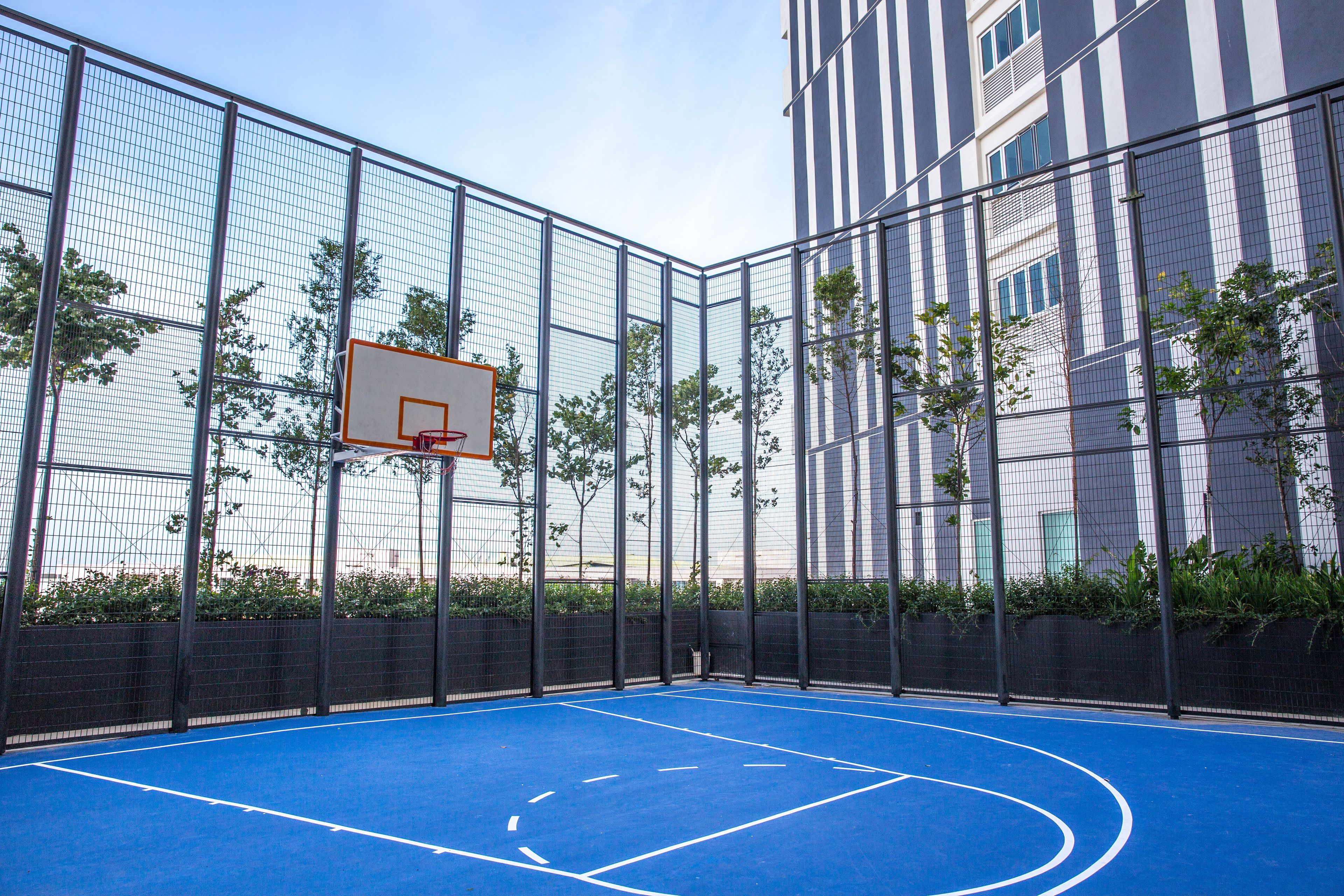 Una cancha de baloncesto al aire libre con aro, rodeada por una cerca metálica y contigua a un edificio con ventanas.