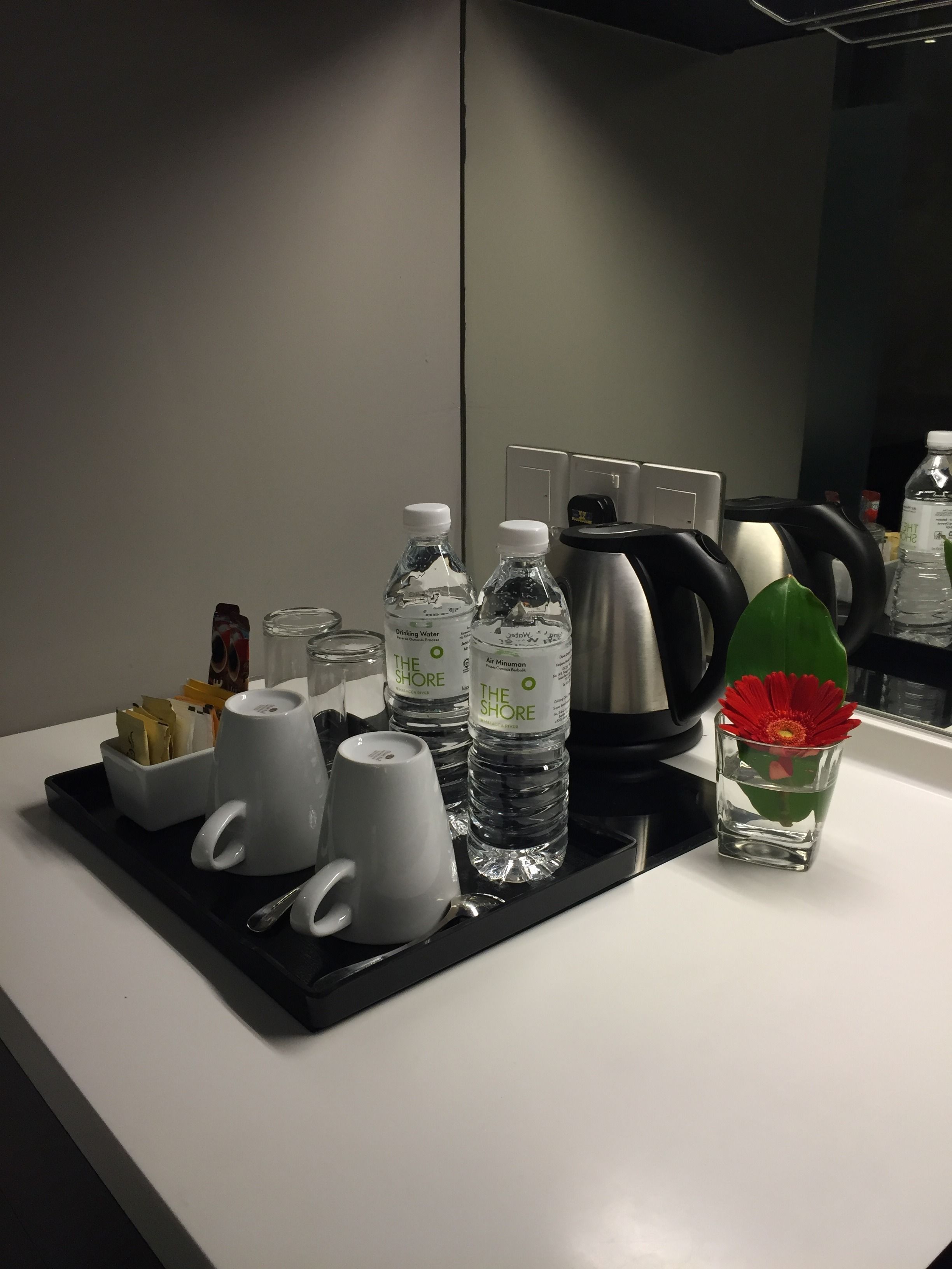 A hotel room counter with a tray holding a coffee maker, two water bottles, a mug, a cup, and a small vase with a flower.