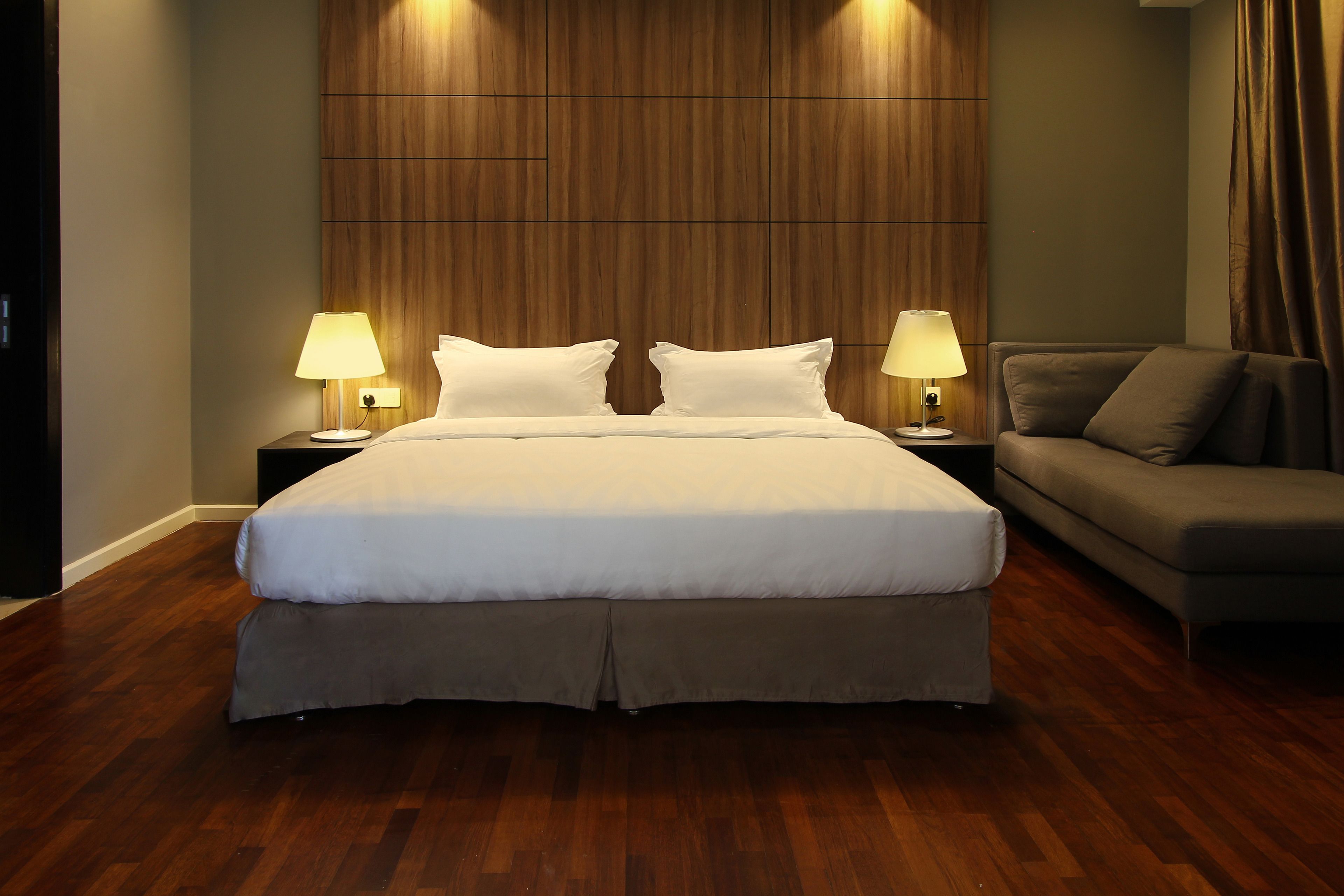 Un dormitorio ordenado con una cama grande, dos mesitas de noche con lámparas, un sofá gris y paneles de madera en la pared.