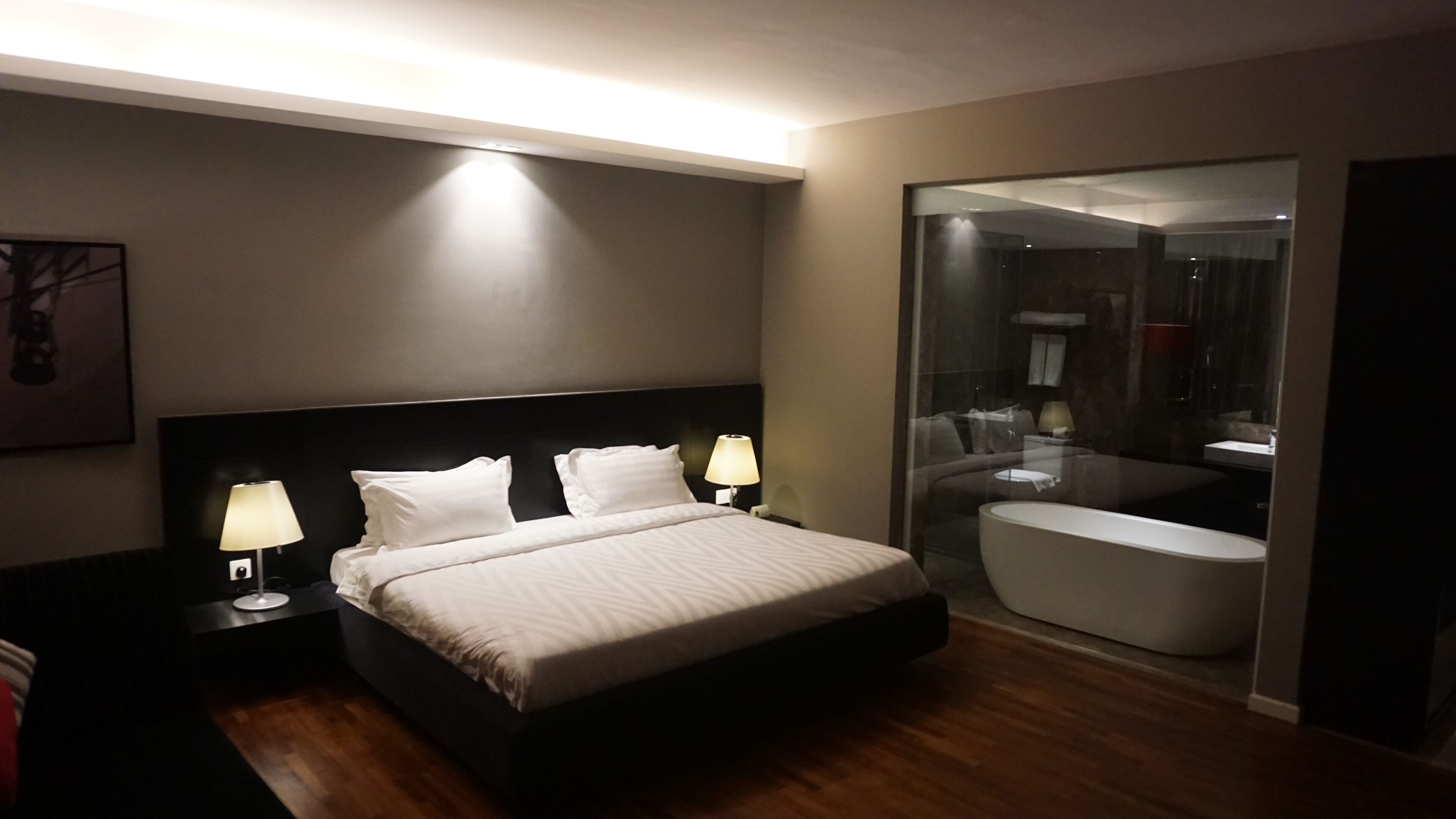 A modern hotel room with a large bed, bedside lamps, a glass-enclosed bathroom with a bathtub, and a framed picture on the wall.