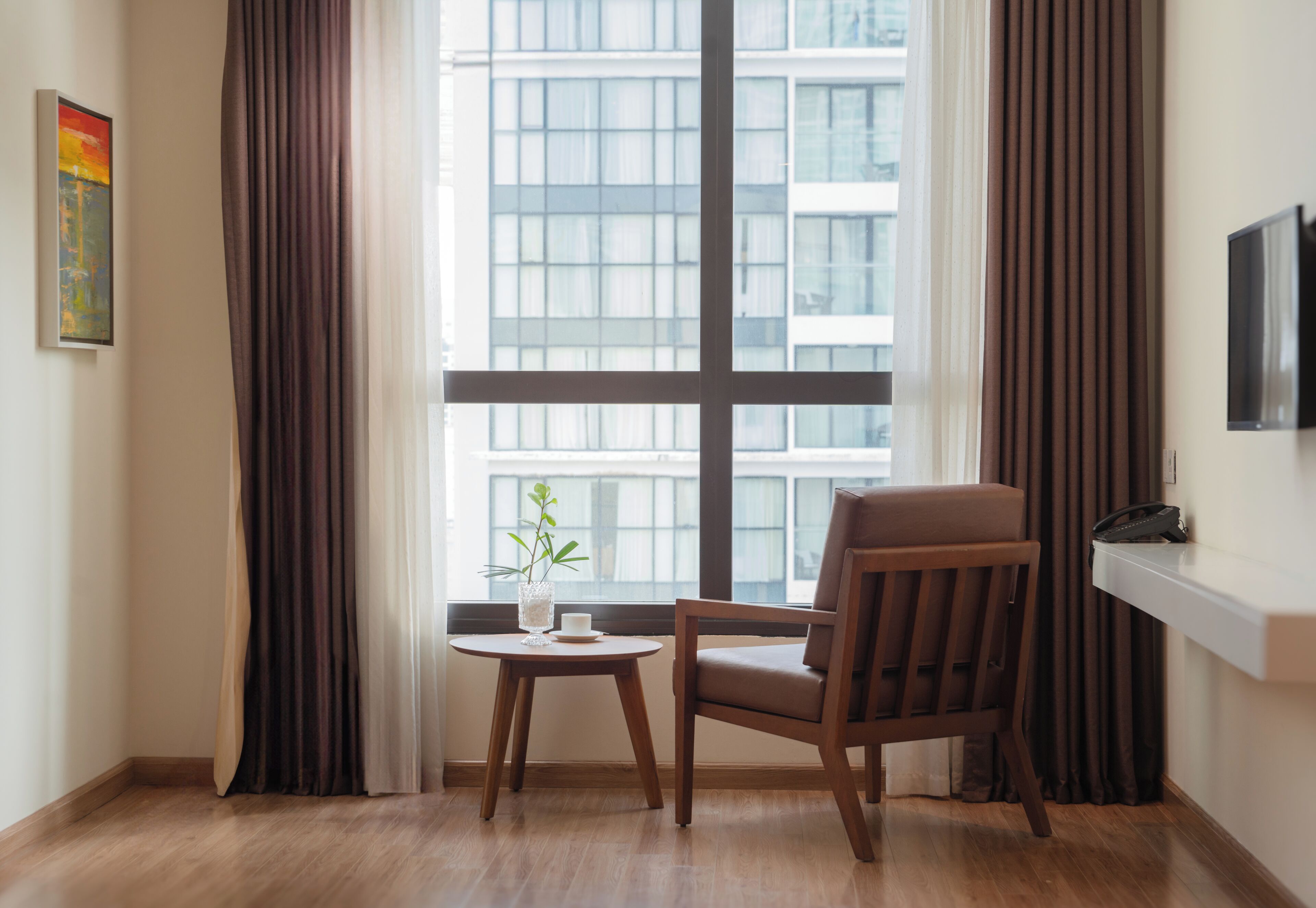 A room with a large window, a chair, a small table with a vase, and a painting on the wall.
