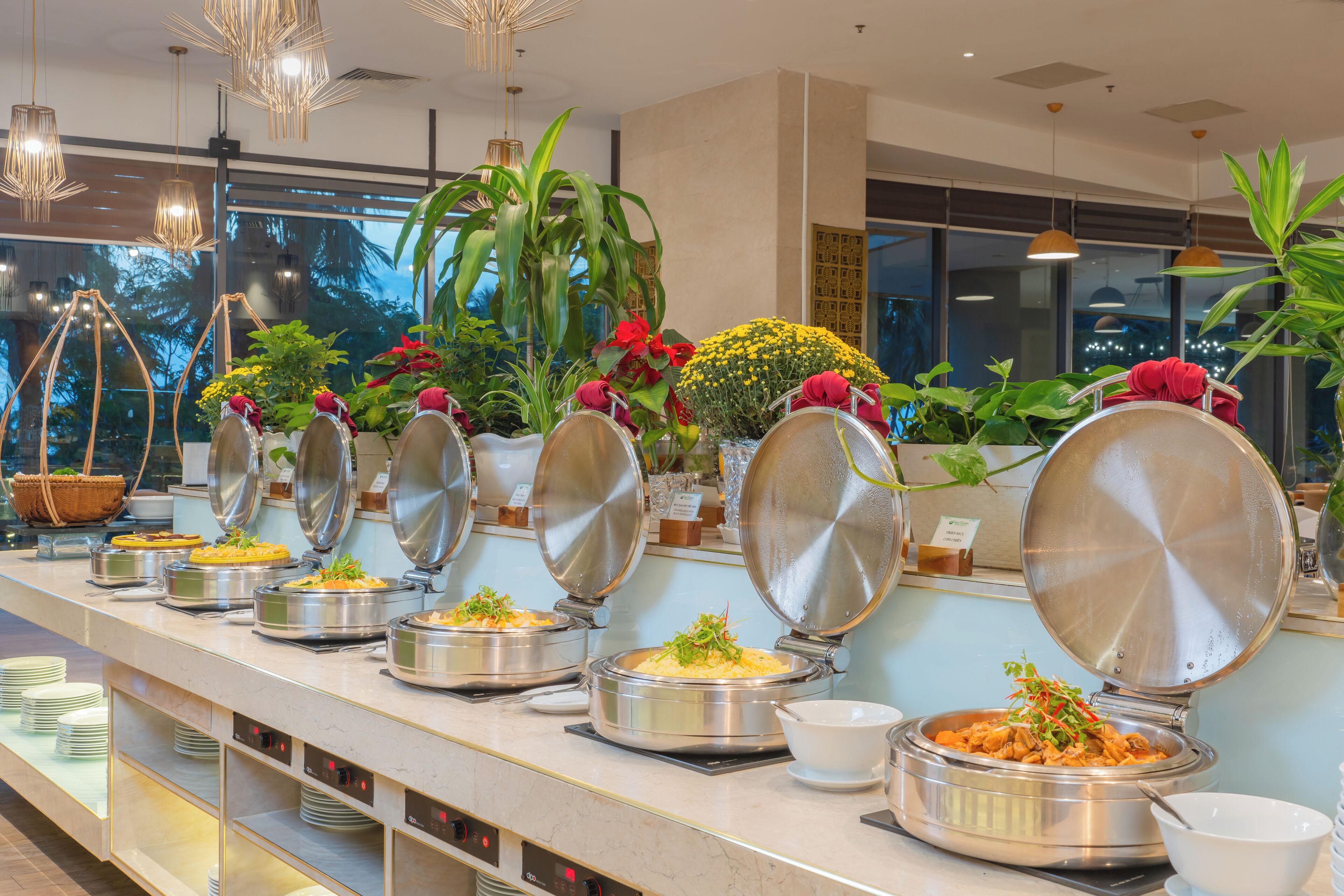 A buffet with various dishes, including a salad, rice, and a noodle dish, with a view of the outdoors through large windows.