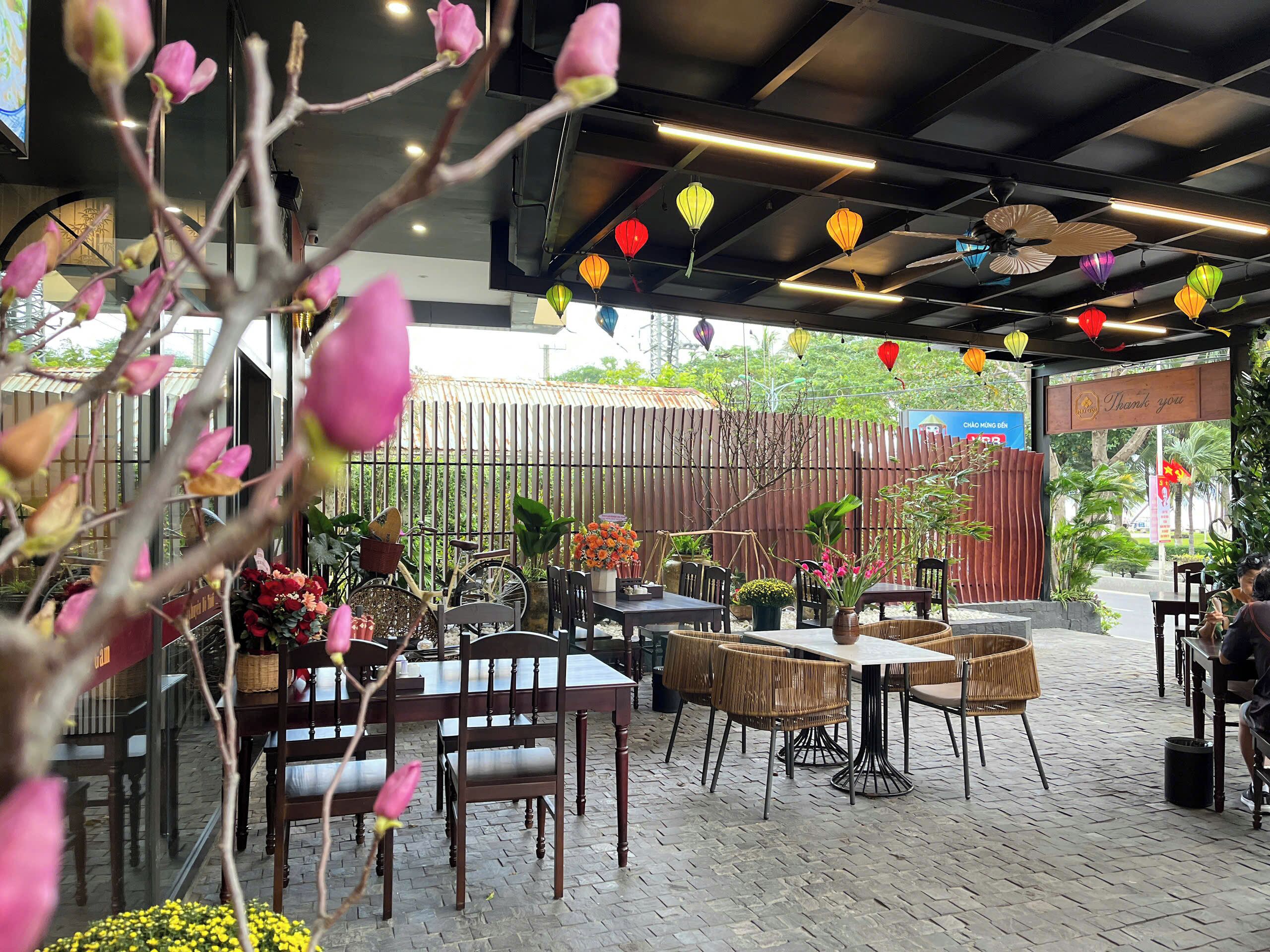 An outdoor café with wooden tables and chairs, surrounded by potted plants and a bamboo fence.