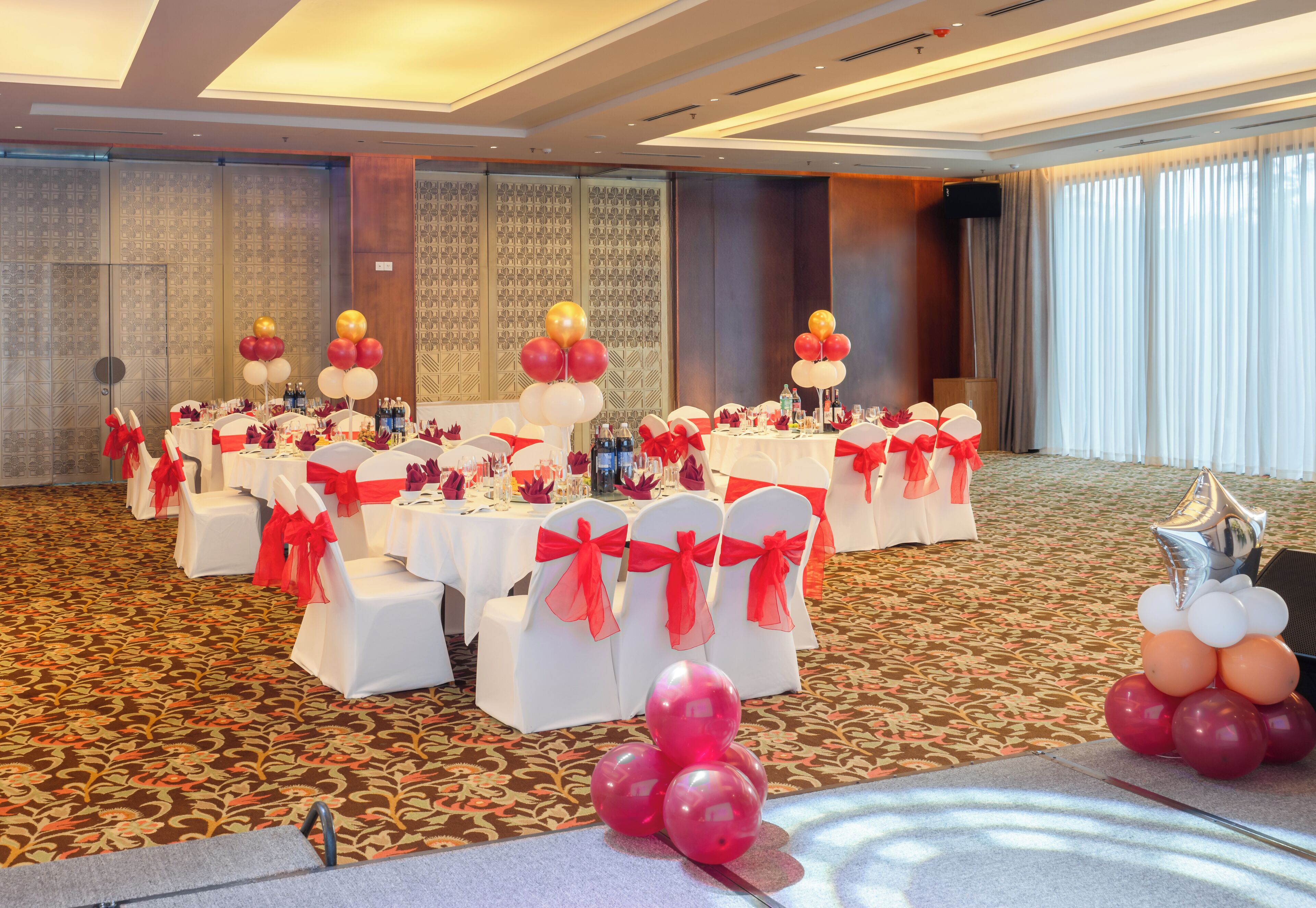 A banquet hall with round tables, white chairs with red sashes, and a patterned carpet.