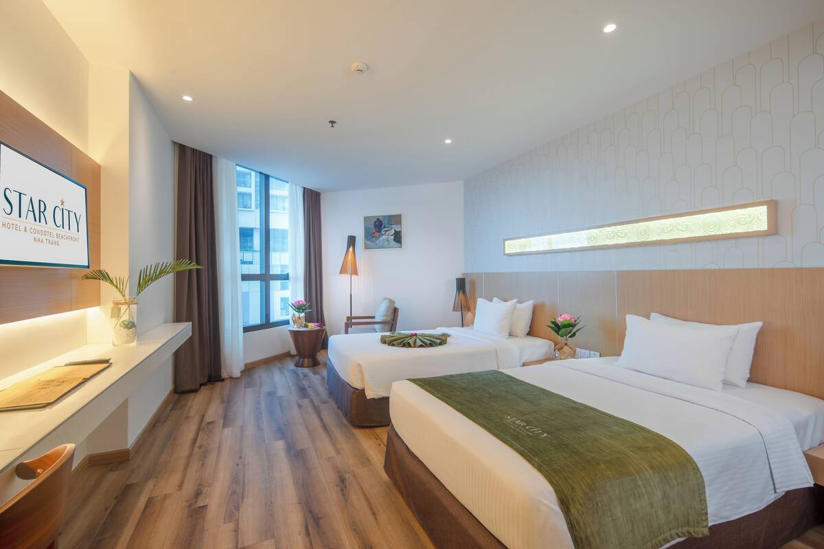 Une chambre d’hôtel avec deux lits, un bureau avec une chaise, une petite table avec un vase et une grande fenêtre avec des rideaux.