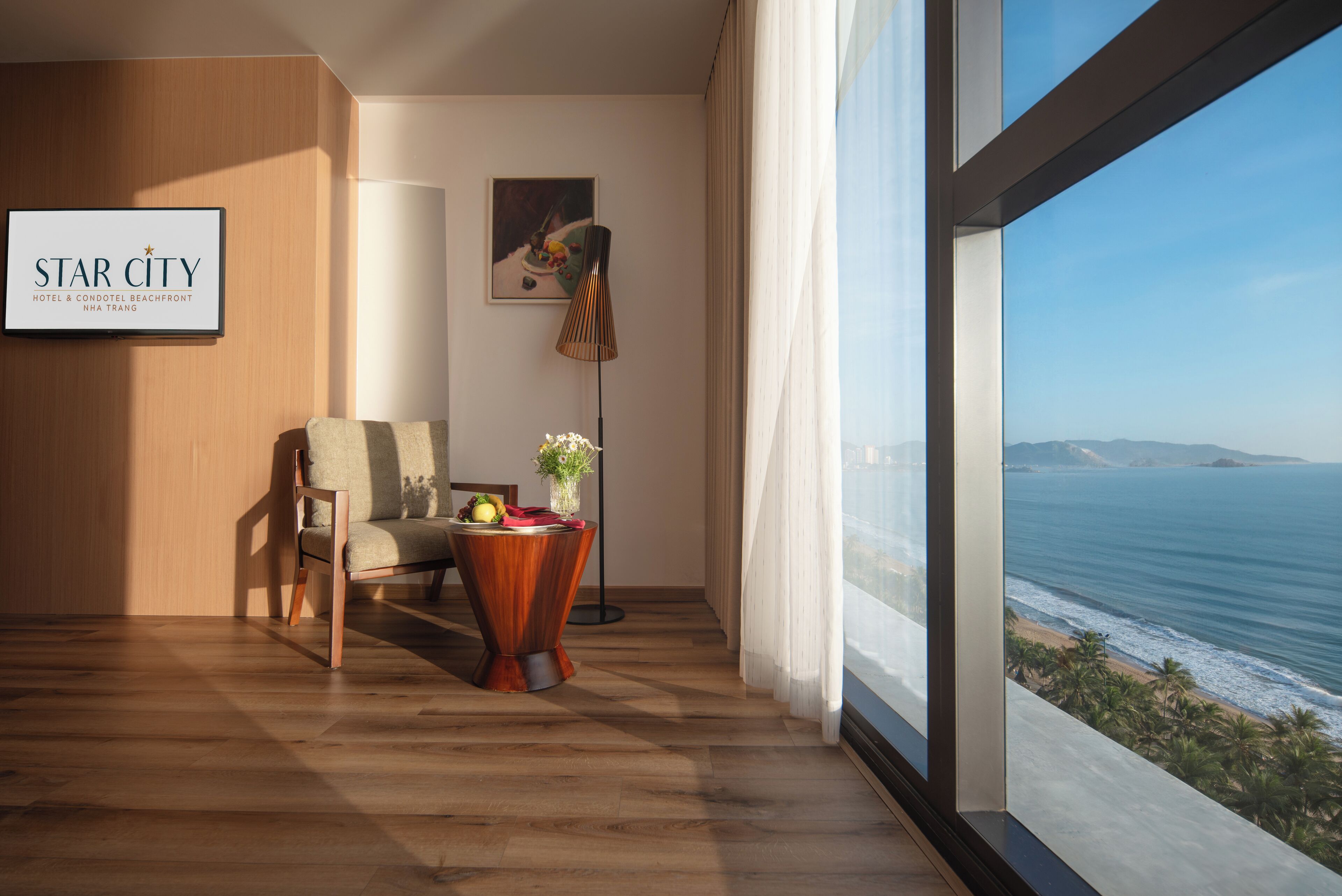 Habitación de hotel moderna con piso de madera, un sillón cómodo, una mesita con un florero y fruta, una lámpara de pie y un amplio ventanal con vistas al océano y la playa.
