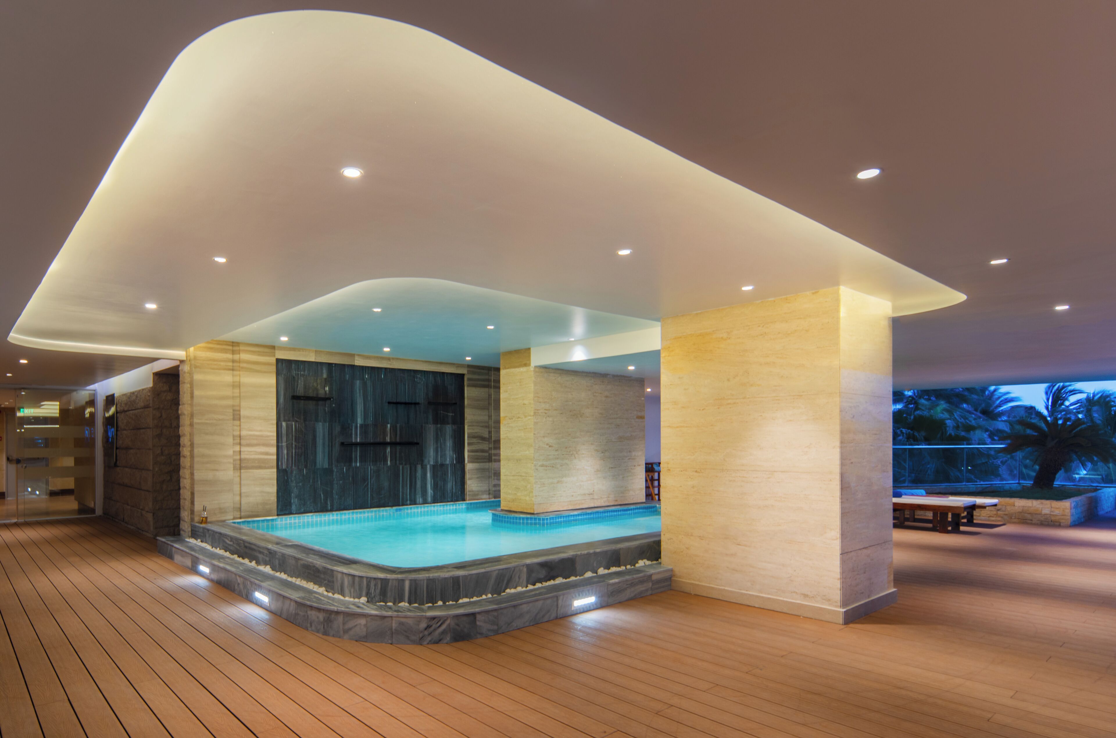 A modern indoor swimming pool with a wooden deck, a curved ceiling, and a stone wall.