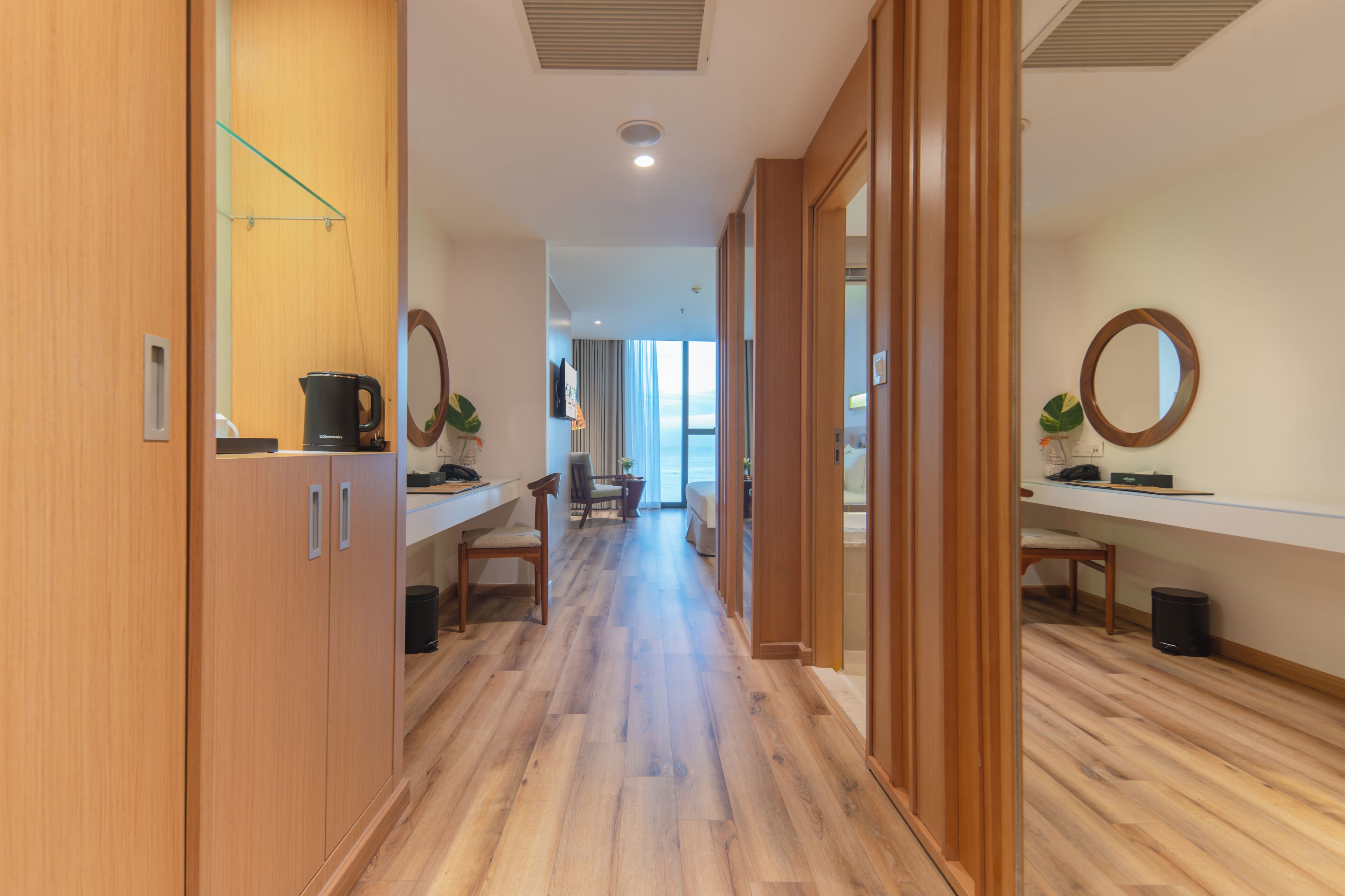 Un couloir d’hôtel moderne avec un plancher en bois, un coin salon avec une table et des chaises, et une vue sur l’extérieur grâce à de grandes fenêtres.