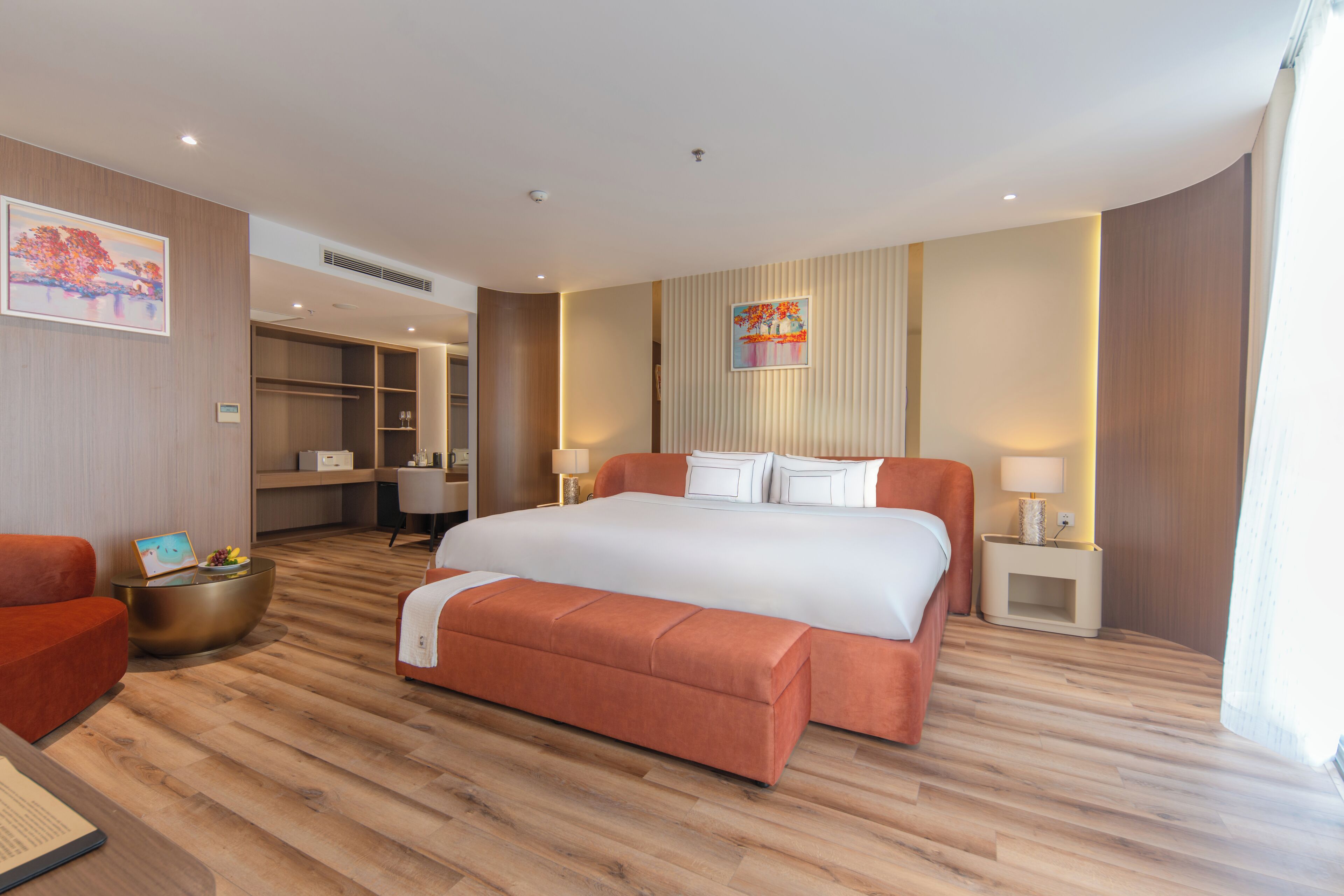 Ein modernes Hotelzimmer mit einem großen Bett, einer Sitzecke mit Sofa und Sesseln, einem kleinen Tisch mit Obstschale und einem Nachttisch mit Lampe.