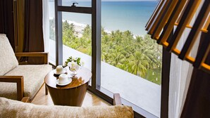 Een kamer met een groot raam dat uitkijkt op een strand en palmbomen, een ronde houten tafel met een bloevas, en een beige bank.