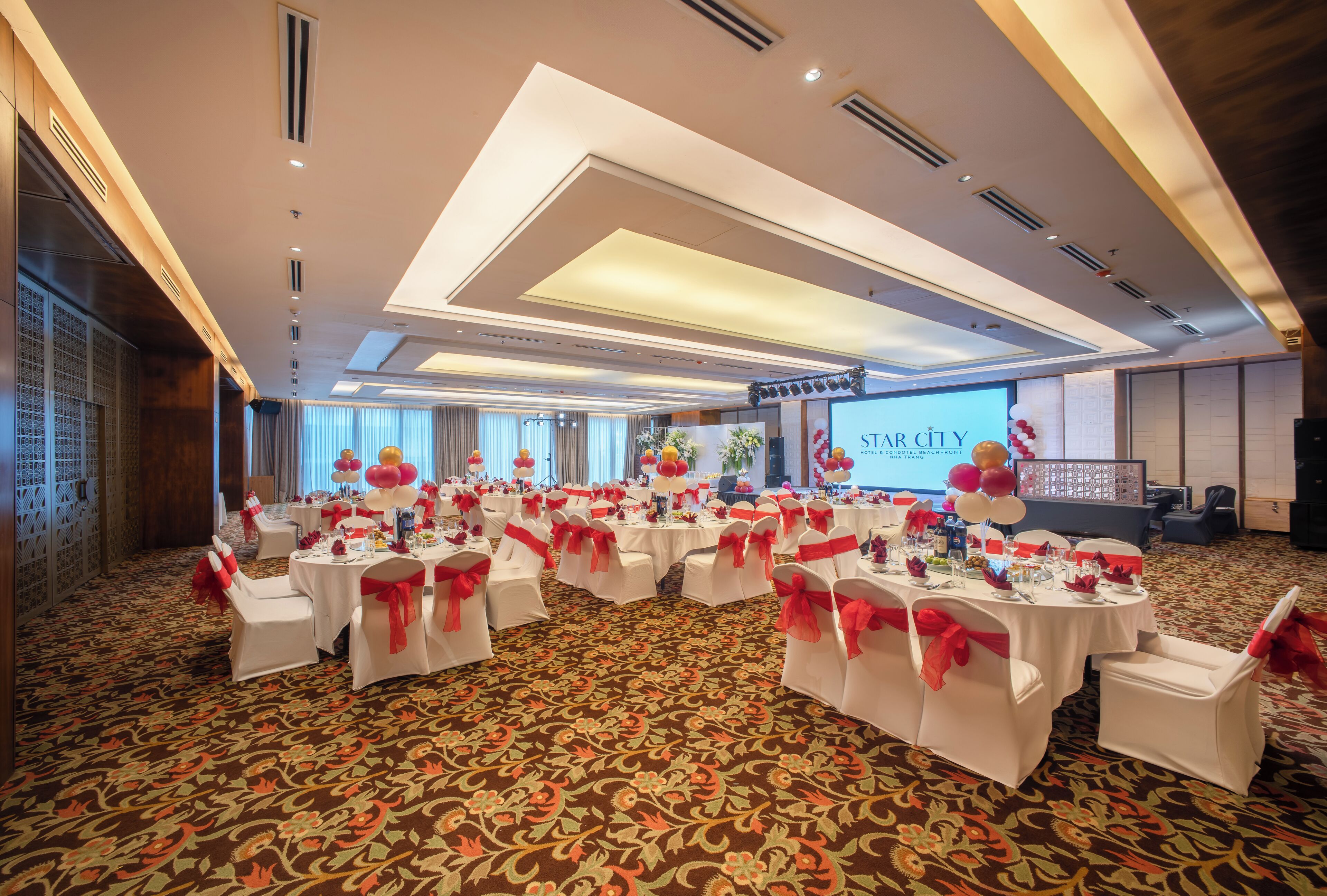 A conference room with multiple round tables, each with white tablecloths and red sashes, set up for a meeting or event.