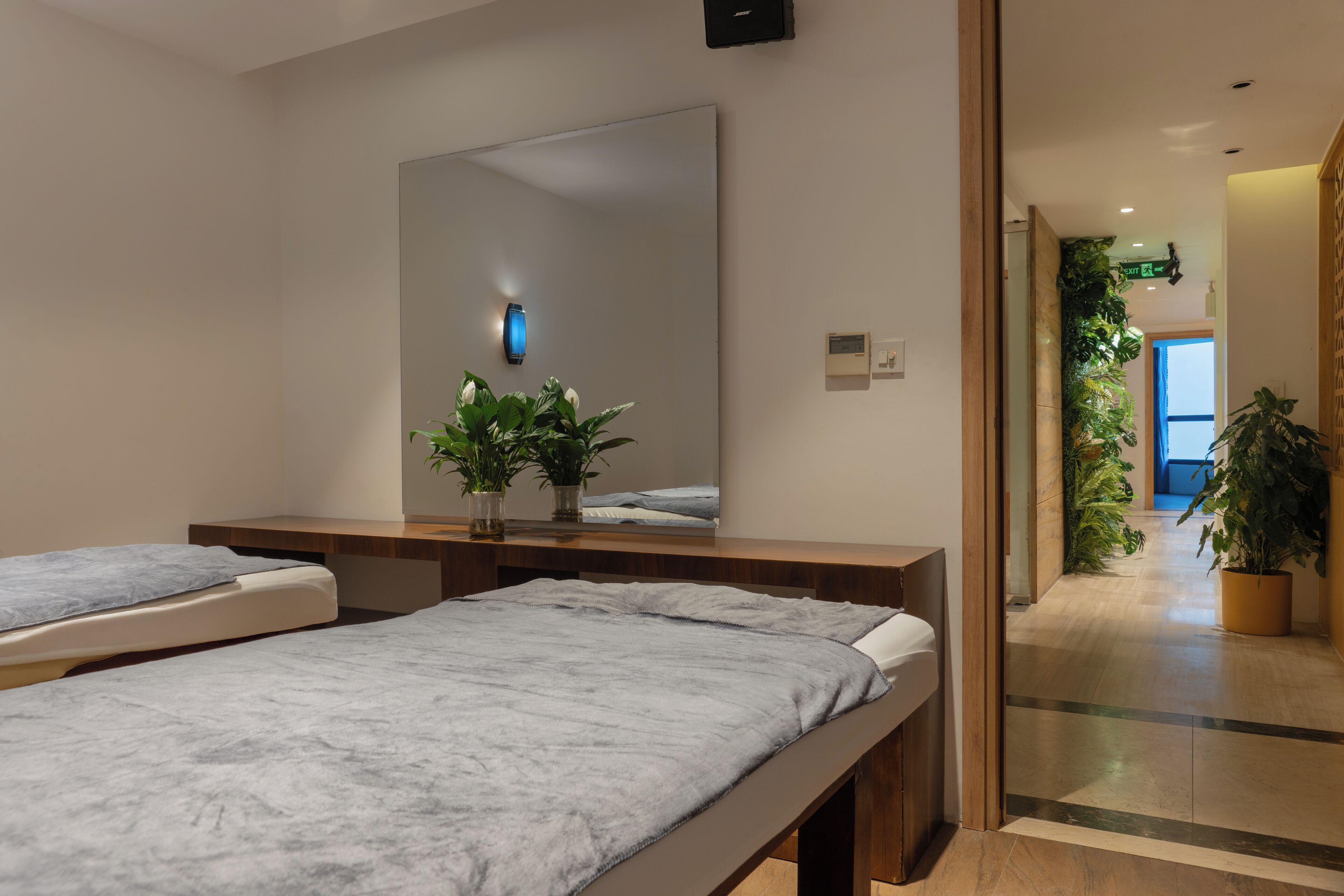 Habitación de hotel con una cama grande, una mesita de noche con una planta, un espejo y vistas a un pasillo con plantas y una puerta.
