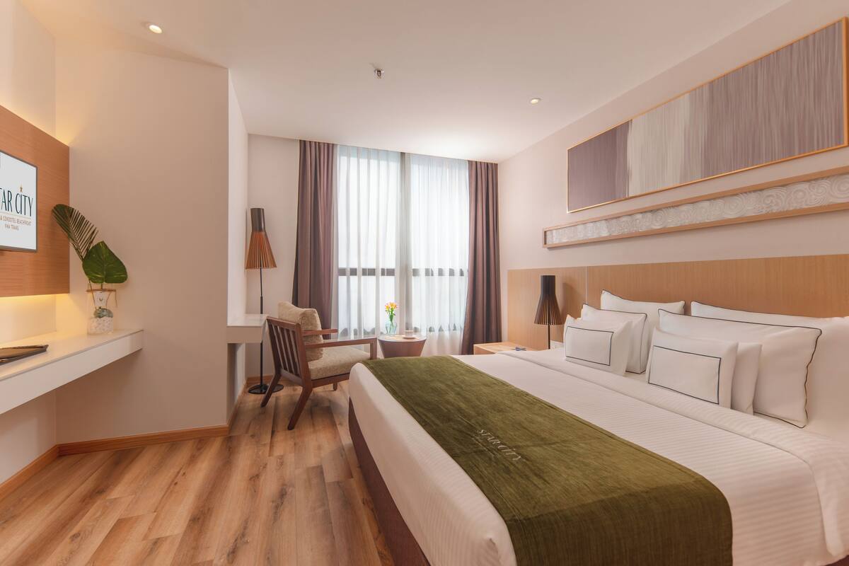 Une chambre d’hôtel avec un grand lit, un bureau, une chaise, une petite table avec un vase et une fenêtre avec des rideaux.