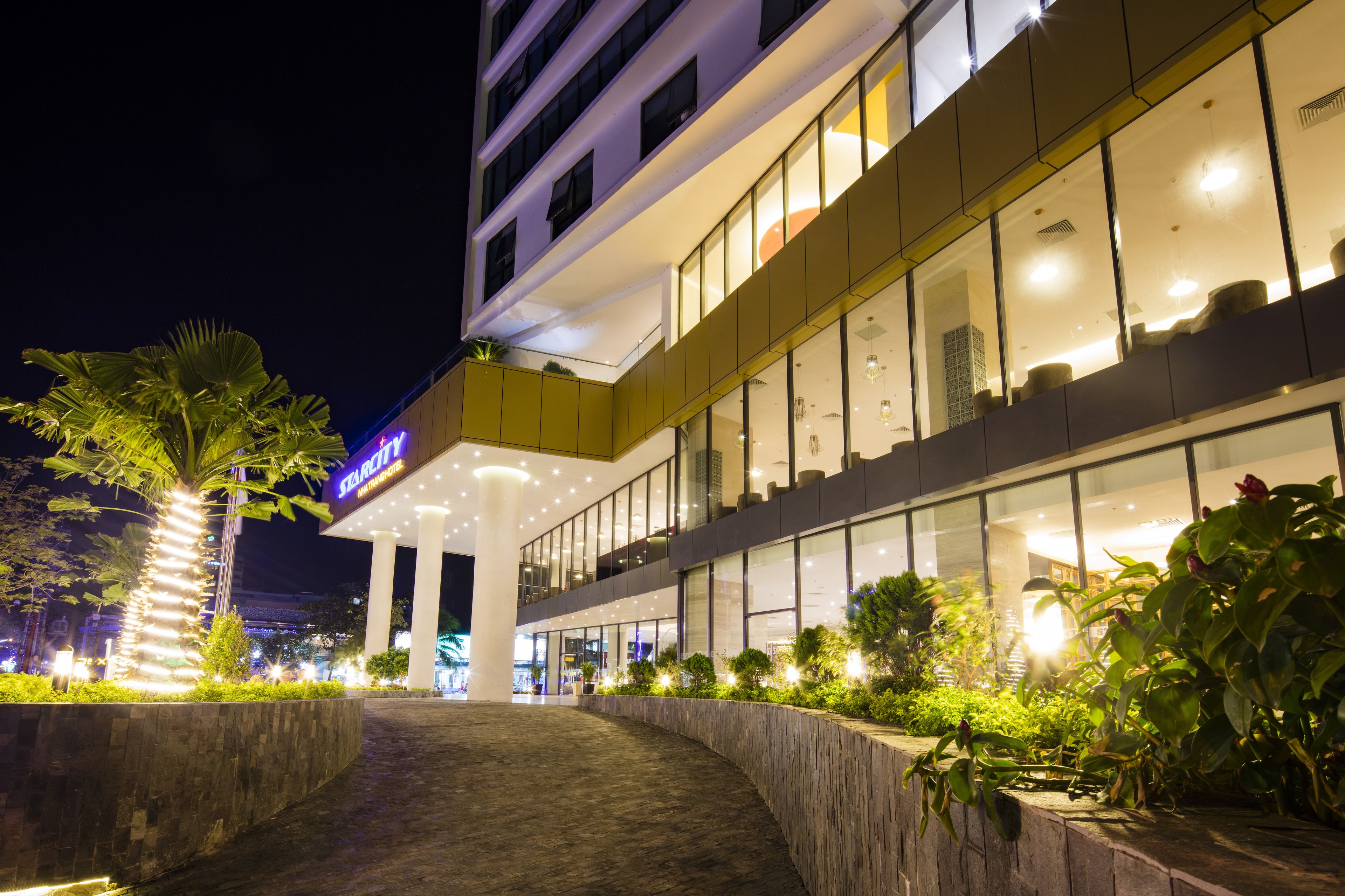 A modern hotel with a well-lit entrance, large windows, and a landscaped area with plants.