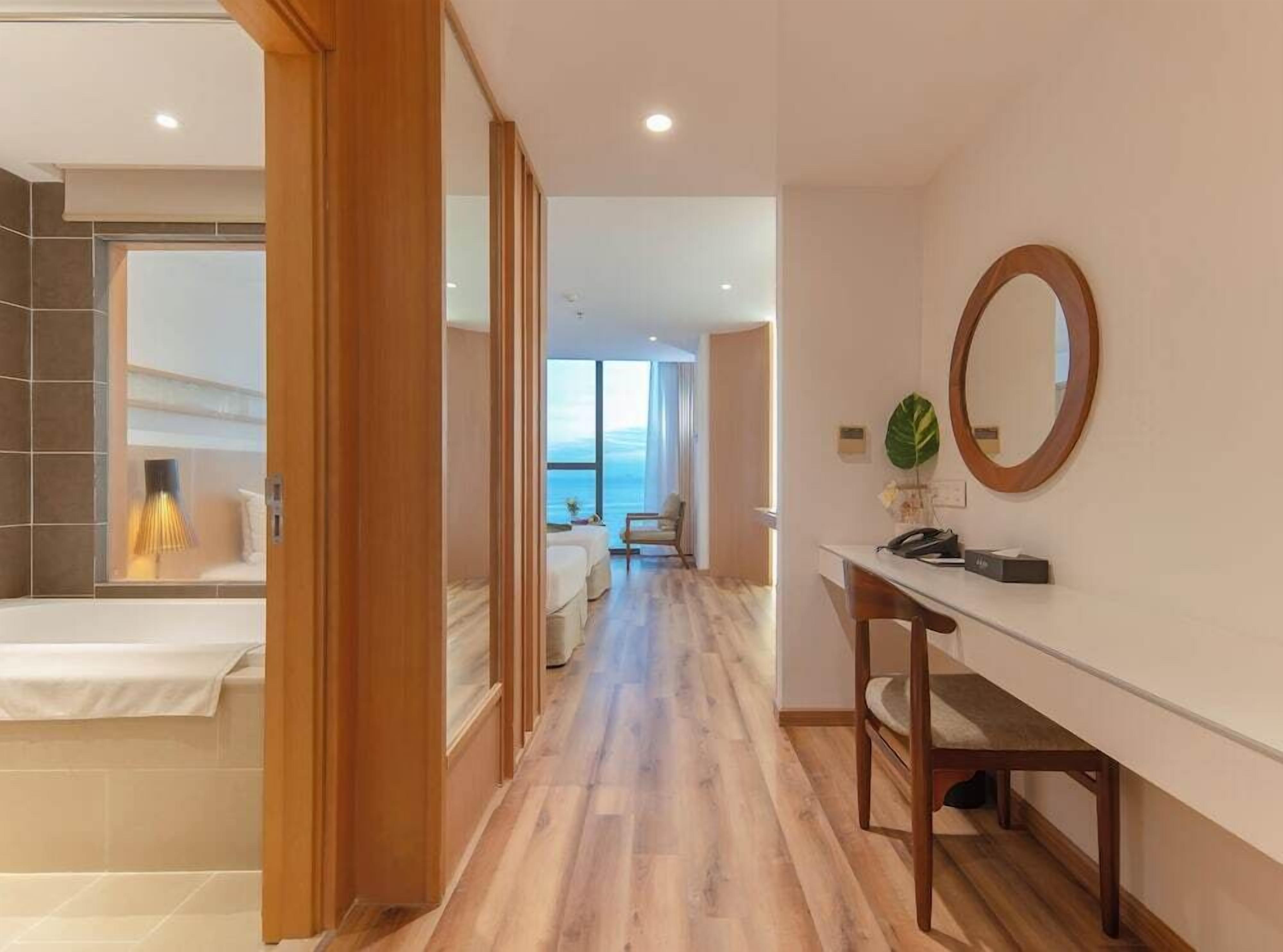 Une chambre d’hôtel avec un plancher en bois, un lit, un bureau avec une chaise, un grand miroir et une vue sur la mer.