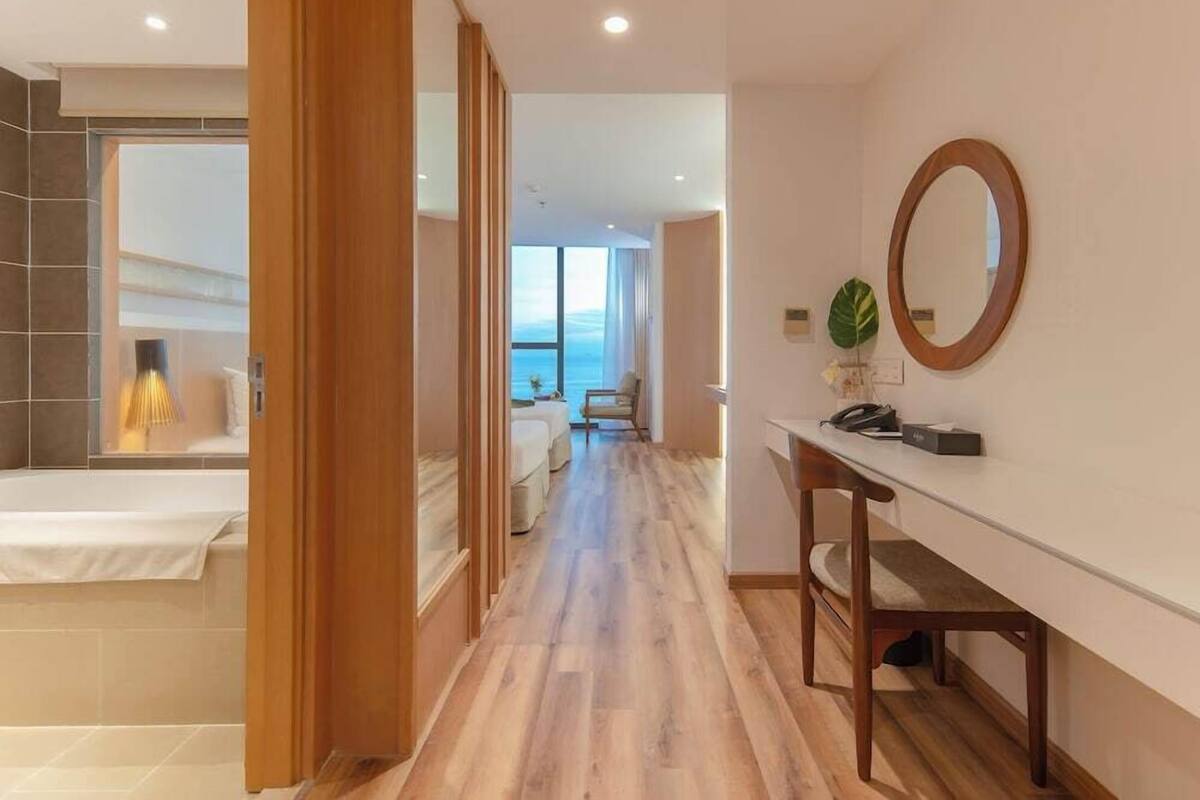 Une chambre d’hôtel avec un plancher en bois, un lit, un bureau avec une chaise, un grand miroir et une vue sur la mer.
