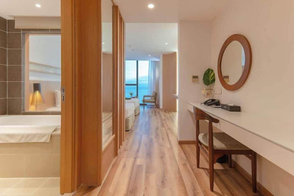Une chambre d’hôtel avec un plancher en bois, un lit, un bureau avec une chaise, un grand miroir et une vue sur la mer.