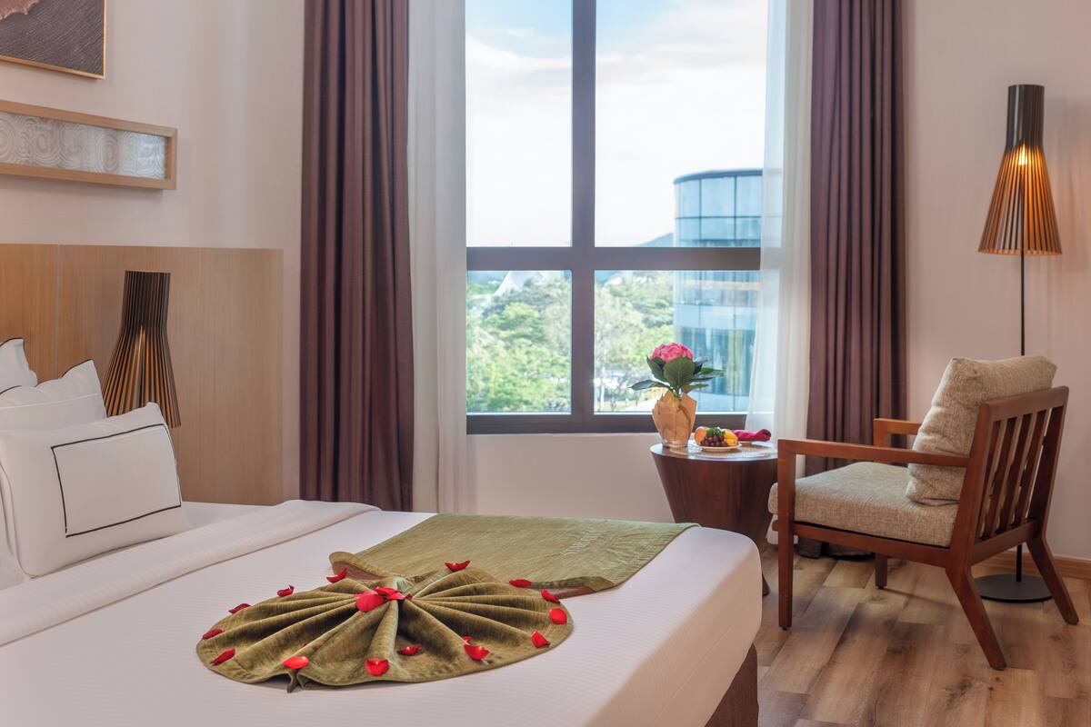 Une chambre d’hôtel avec un lit, une chaise, une table ornée d’un vase de fleurs et une vue sur la verdure environnante.