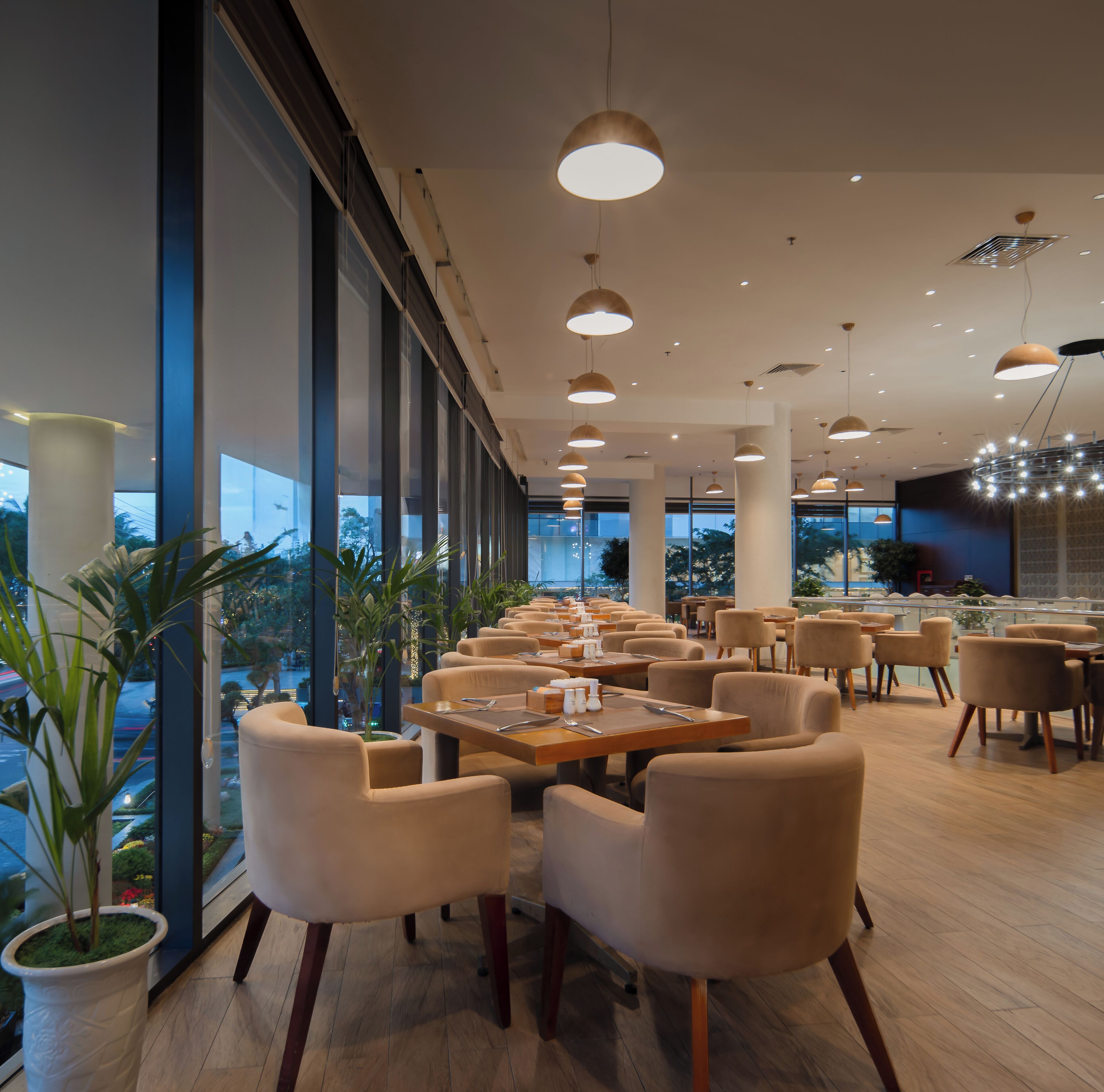 A modern restaurant with high ceilings, pendant lighting, and large windows offering a view of the outdoors.
