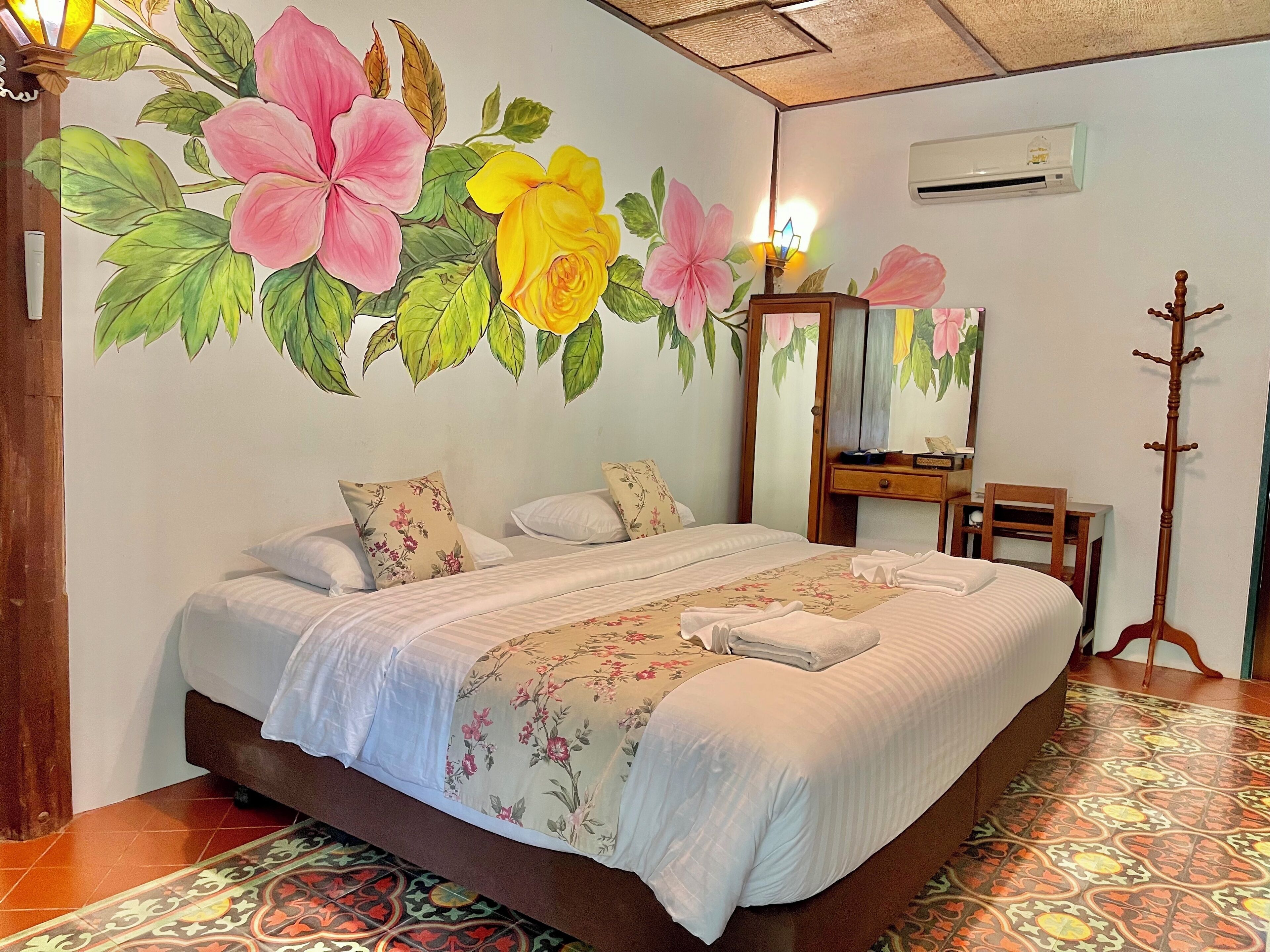 Una habitación de hotel con una cama grande, decoración floral en la pared, una mesita de noche de madera, un espejo y una lámpara de pie.