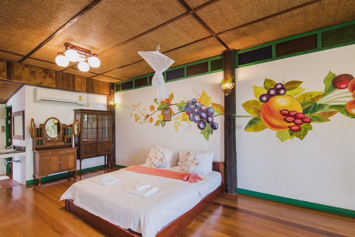Une chambre d’hôtel avec un grand lit, des meubles en bois, une fresque représentant des fruits et un plafond en chaume.
