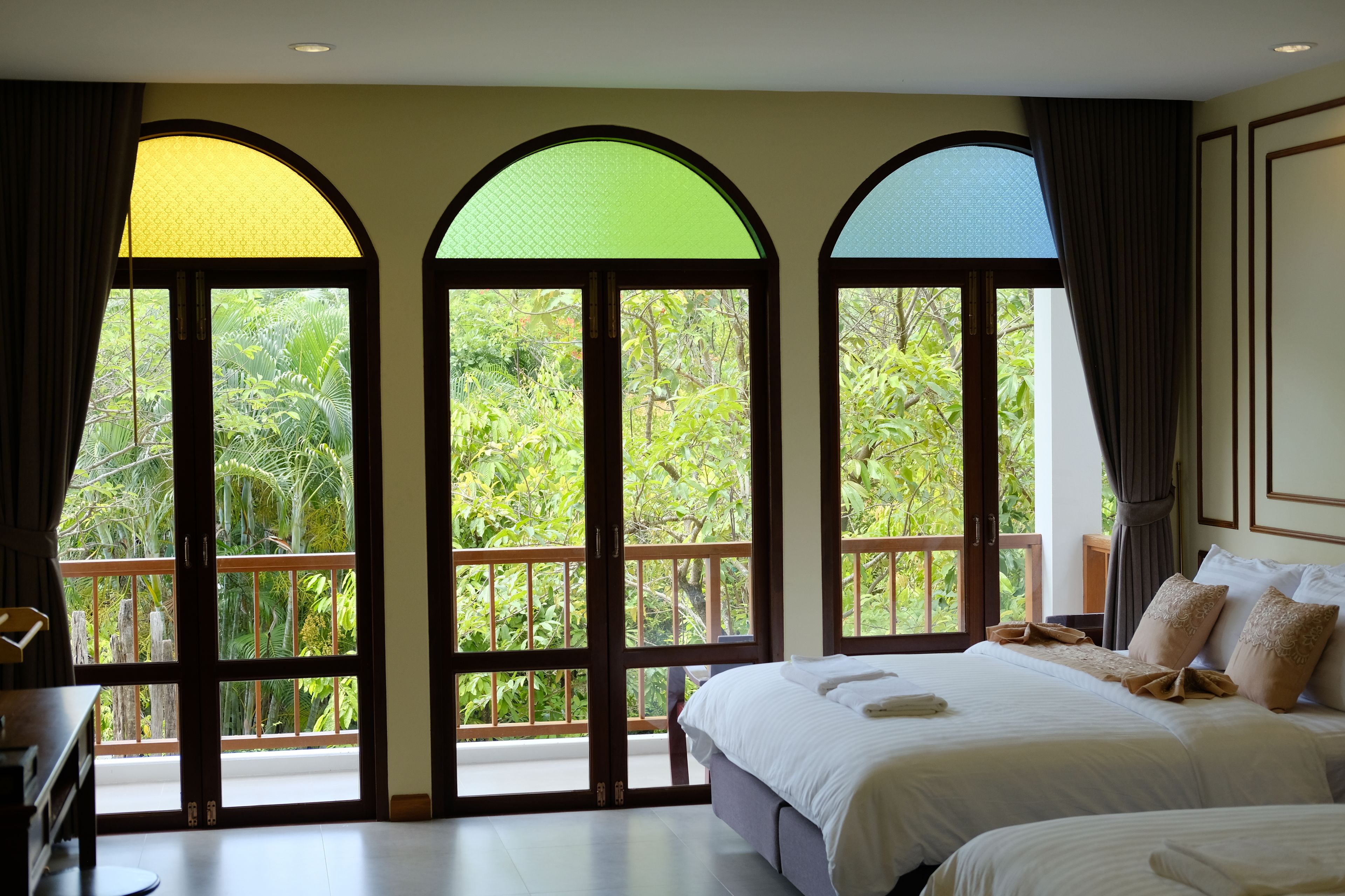 A hotel room with a large bed, a desk, and three arched windows offering a view of lush greenery.
