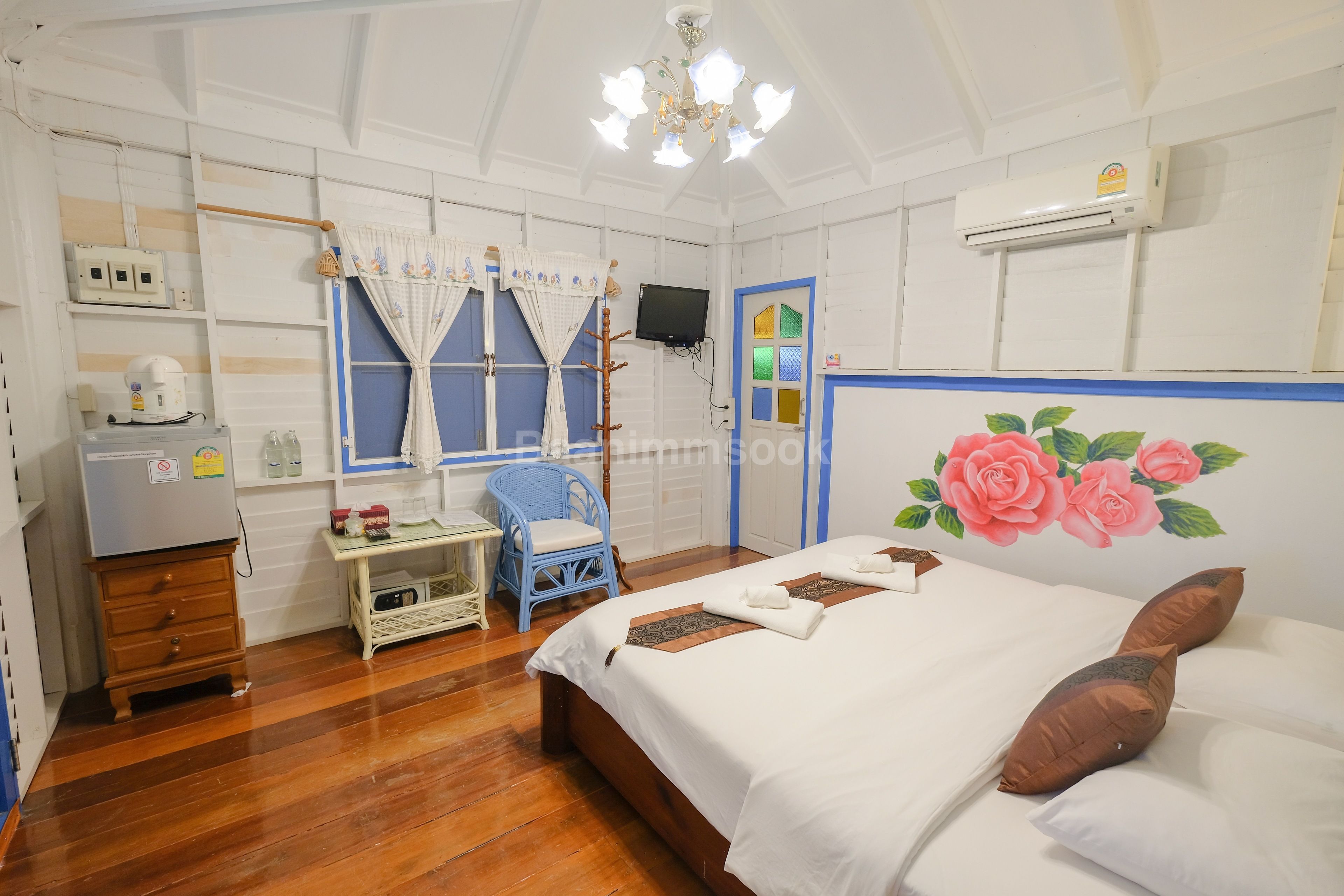 A bedroom with a wooden floor, a bed with white bedding, a blue chair, a small table with a kettle, and a TV on the wall.