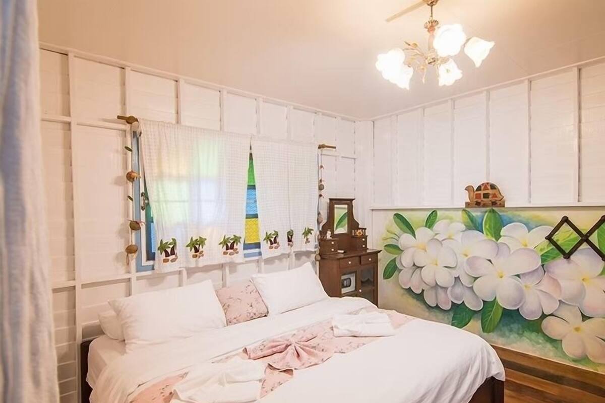 Une chambre à coucher avec un lit, une commode, un miroir et un tableau représentant des fleurs accroché au mur.